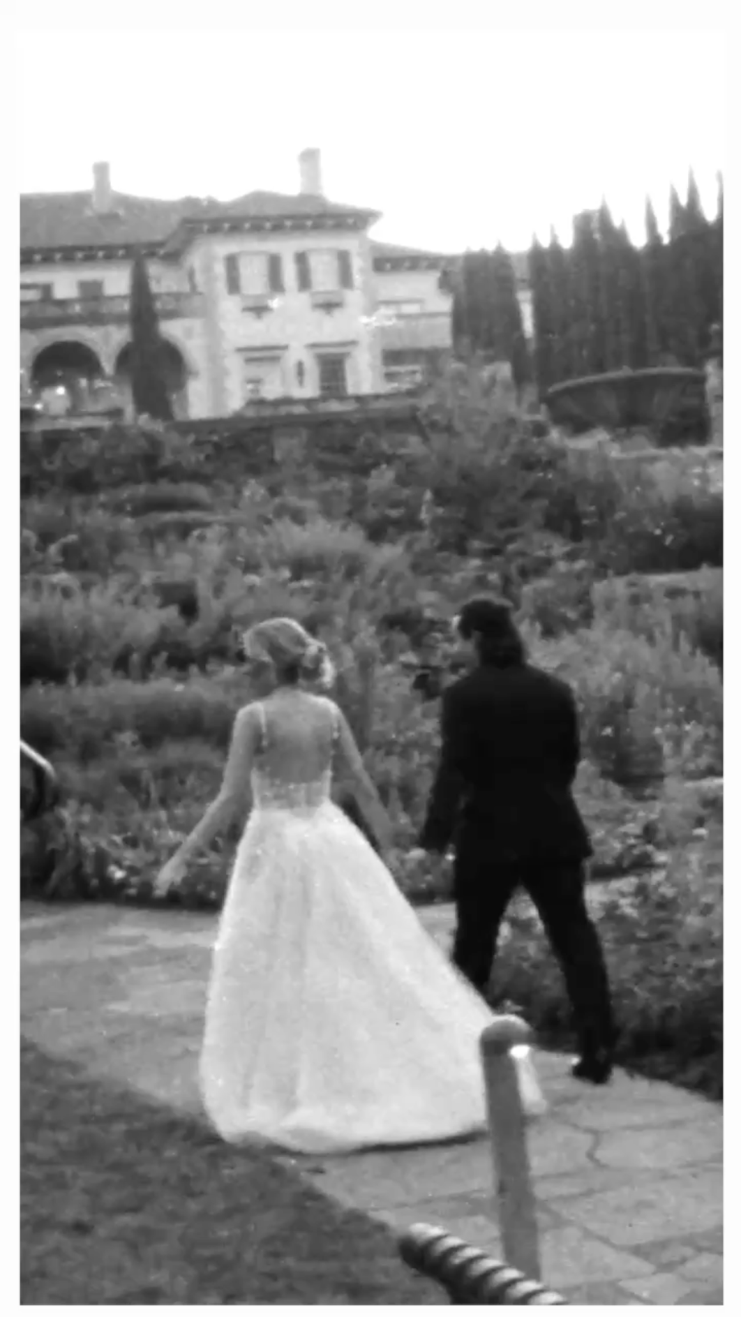 A black and white photo of a bride in a wedding dress and a groom in a tuxedo holding hands and walking outdoors.