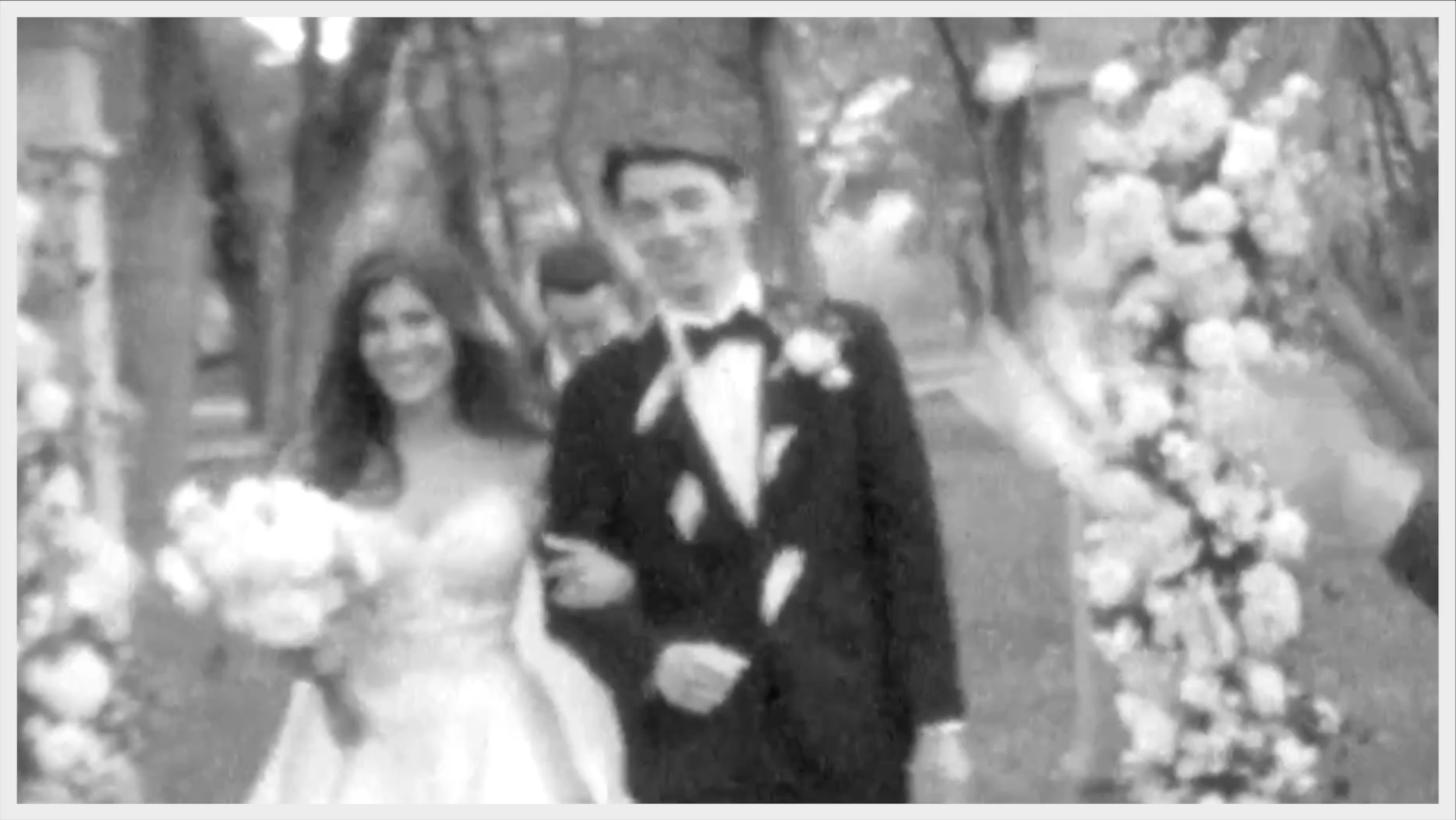 Black and white photo of a bride and groom standing outdoors with floral arch and trees in the background, wedding attire, smiling.