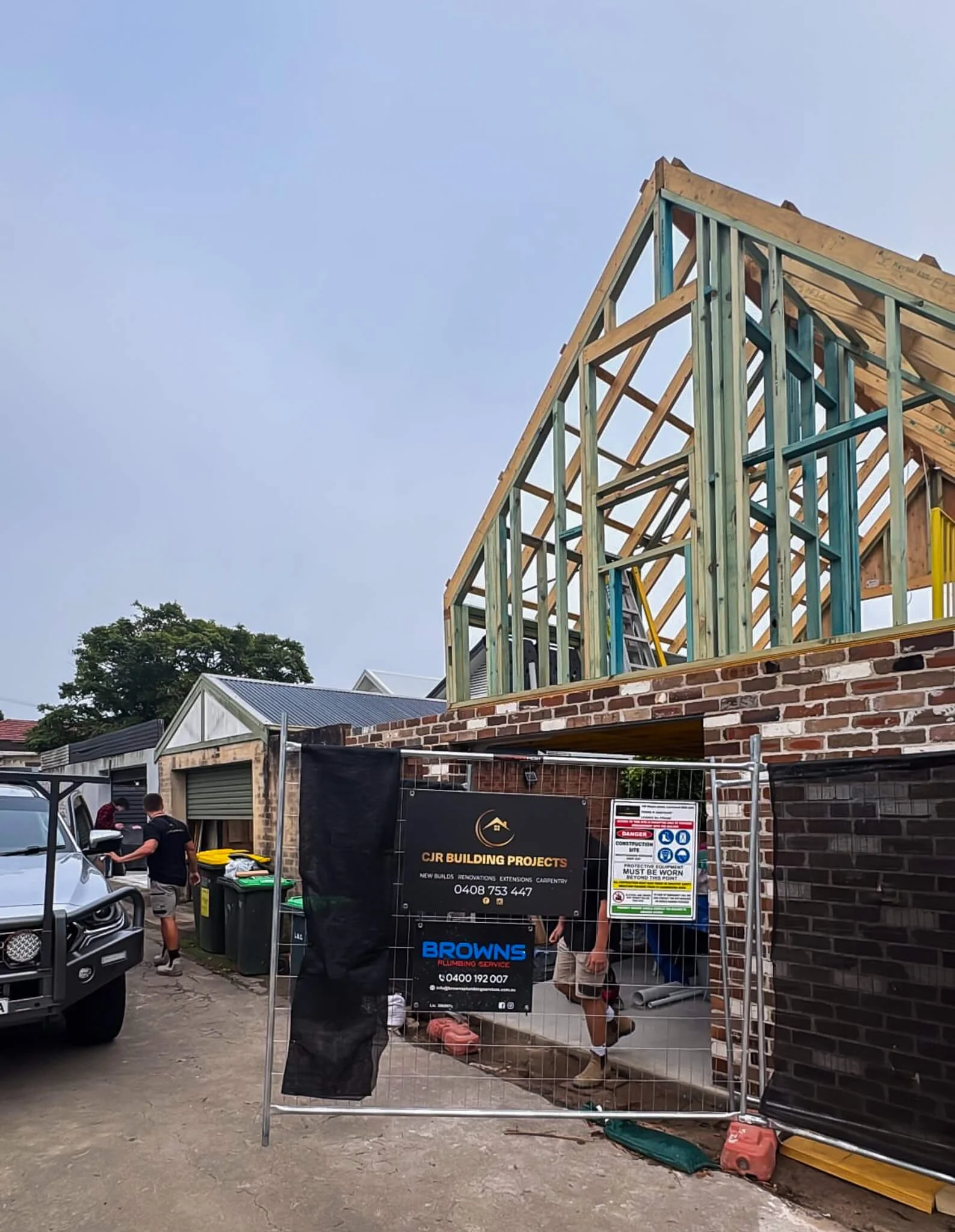 Construction site with house framing in progress, workers on site, construction signs, and partially built brick walls.