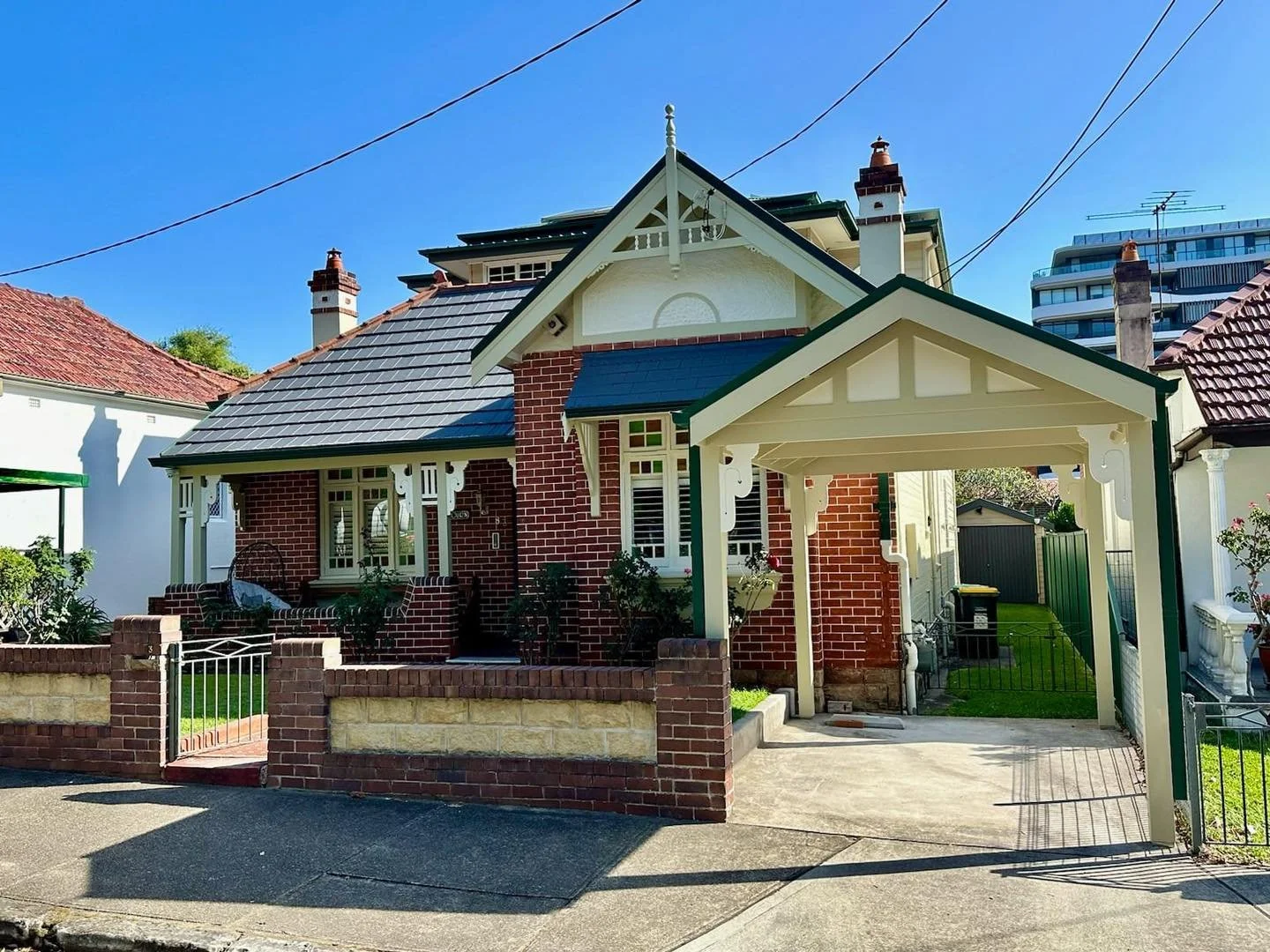 A Victorian-style house with red brick walls, white decorative trim, and a green roof. The house has a front porch with brick steps and a small garden, and a driveway with a covered carport to the right. There are other houses and a modern building in the background against a clear blue sky.