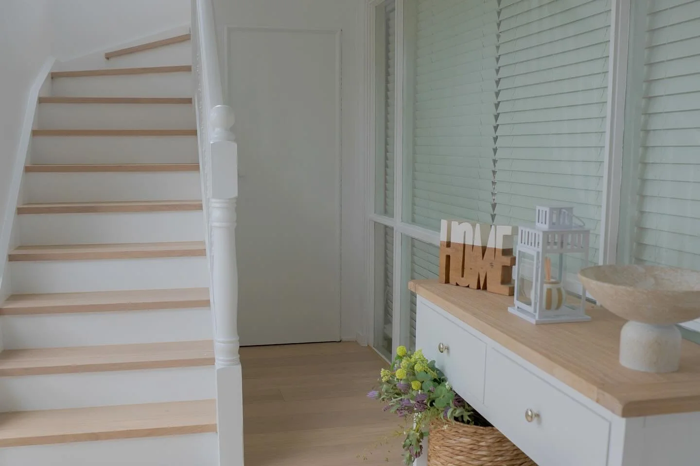 Interior view of a bright, minimalistic home entryway with a staircase on the left, a white sideboard with decorative items on the right, and a window with closed blinds in the background.