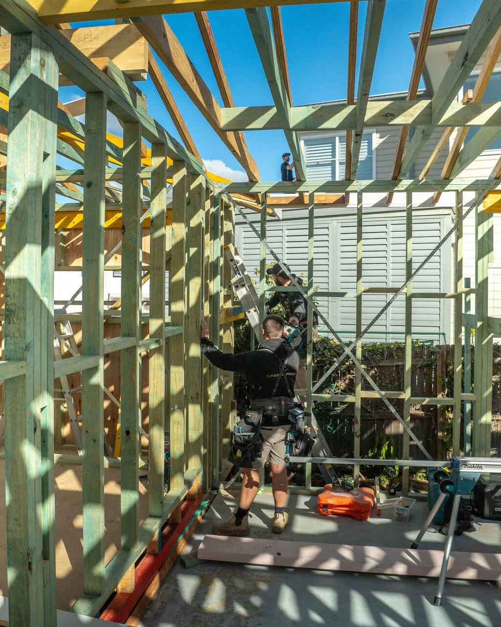 Construction workers building a framed structure in a backyard on a sunny day, with blue skies overhead.