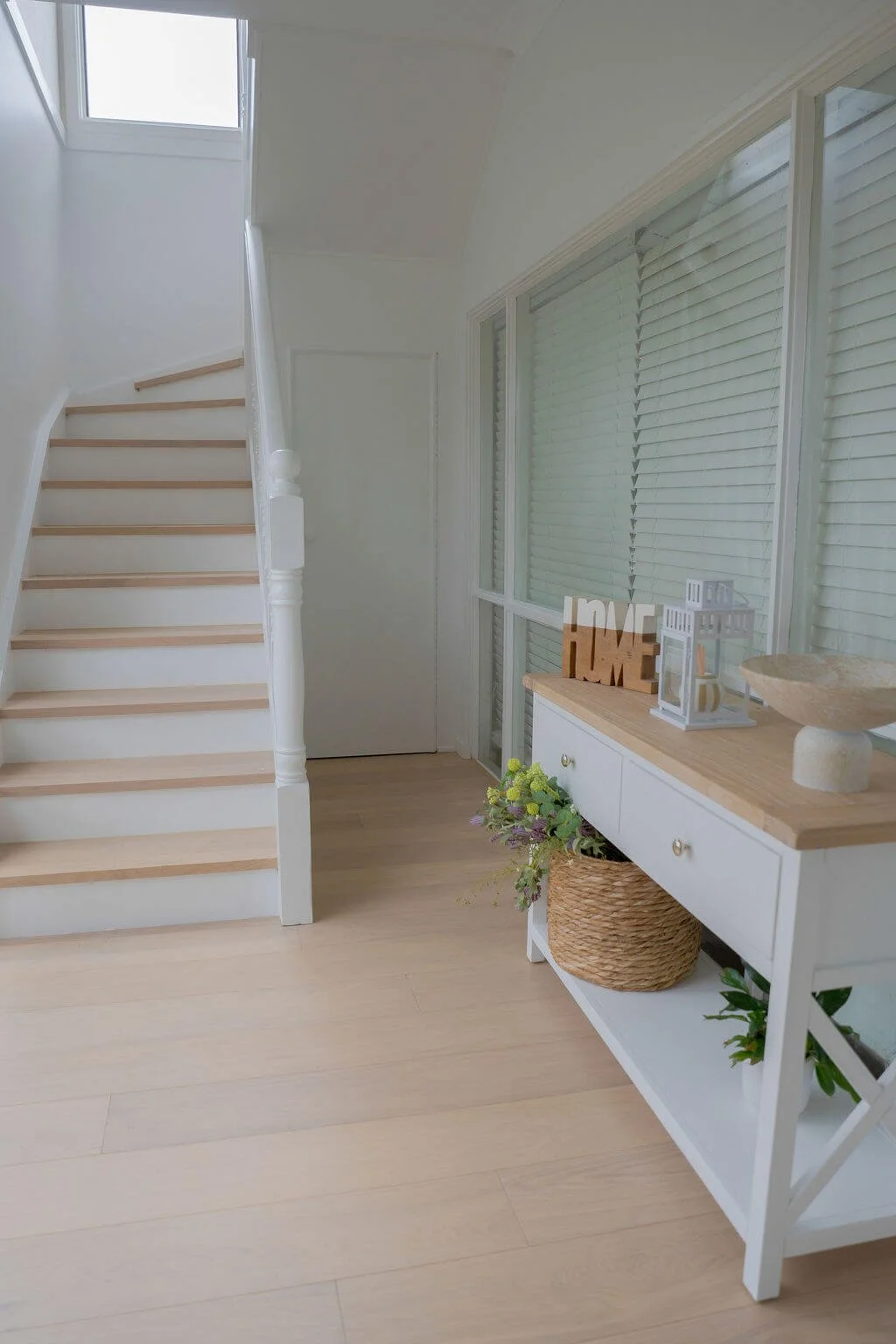 Bright entryway with a white wooden staircase, a skylight, a table with decorative items, a wicker basket with flowers, and large windows with closed blinds.