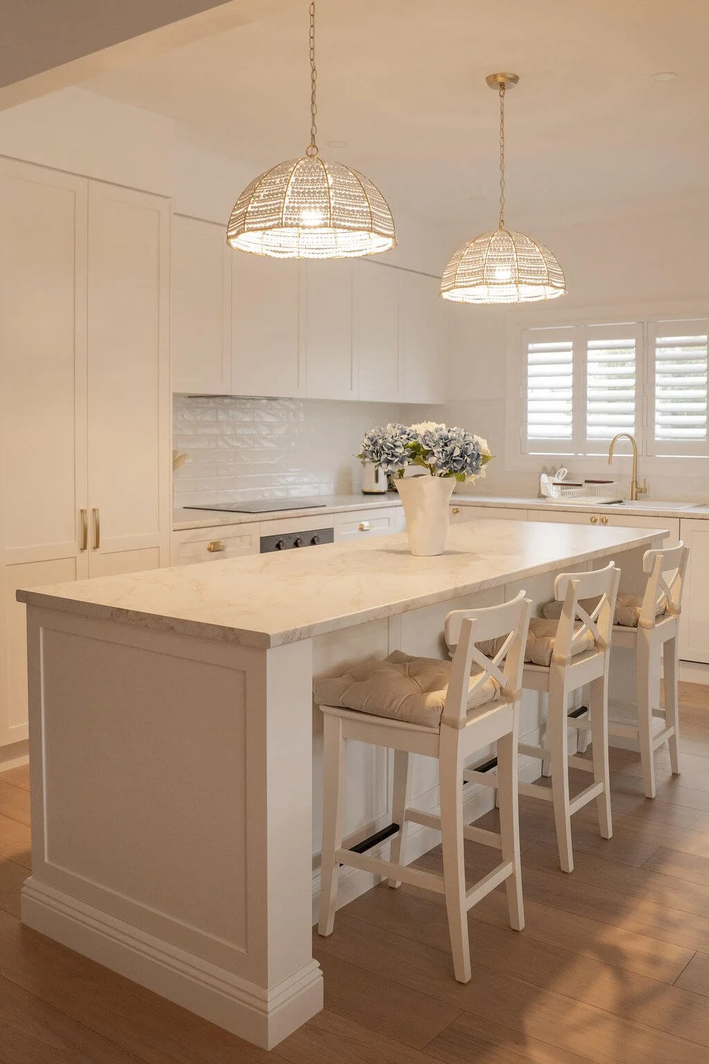 Bright white kitchen with a marble island, three white bar stools with cushions, pendant lights, a white flower vase, and window shutters.