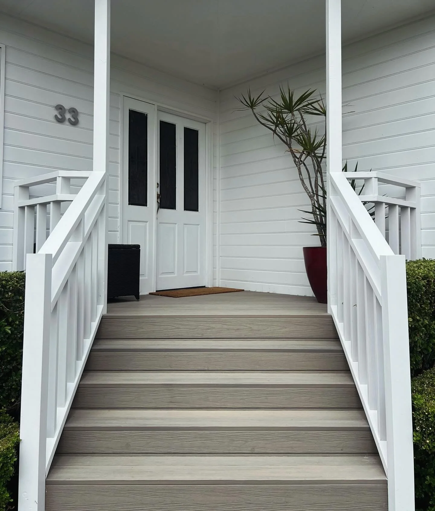 Front porch of a house with a white door, a potted plant on the right, and a black wicker side table on the left. The house number 33 is visible on the wall to the left of the door. There are white railings and gray stairs leading up to the porch.