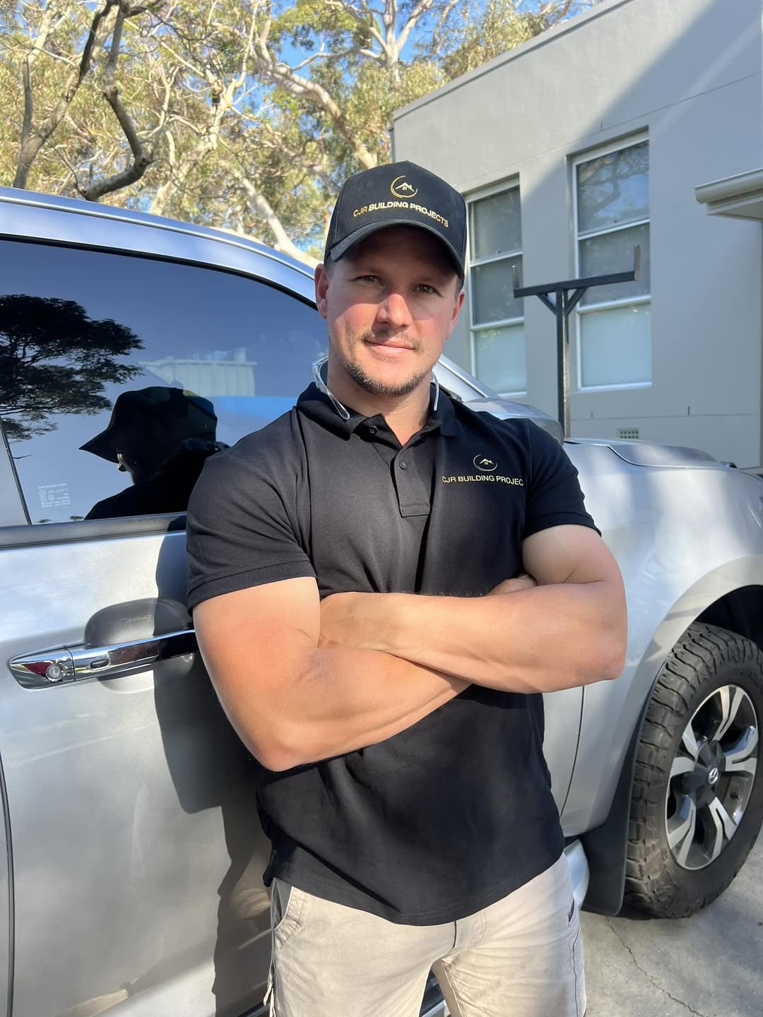 Man standing with arms crossed in front of a silver SUV, wearing a black polo shirt and black cap, both with a logo and the text 'CJR Building Projects,' outdoors with trees and a house in the background.