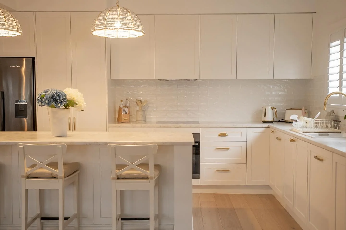 A bright, modern kitchen with white cabinets, countertops, and a backsplash. There is a kitchen island with two white chairs with padded seats. On the island, there is a white vase with blue and white flowers. The kitchen has a stainless steel refrigerator, a toaster, and a kettle, with light wood flooring and a window with blinds.