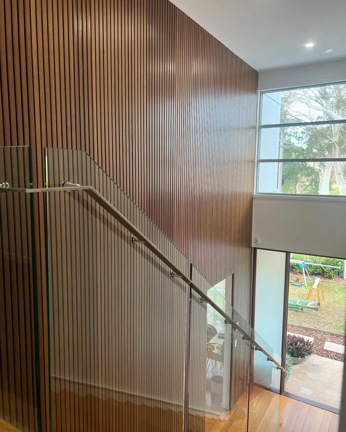 Interior view of a staircase with wooden wall paneling, glass railing, large windows revealing a yard with trees and lawn.