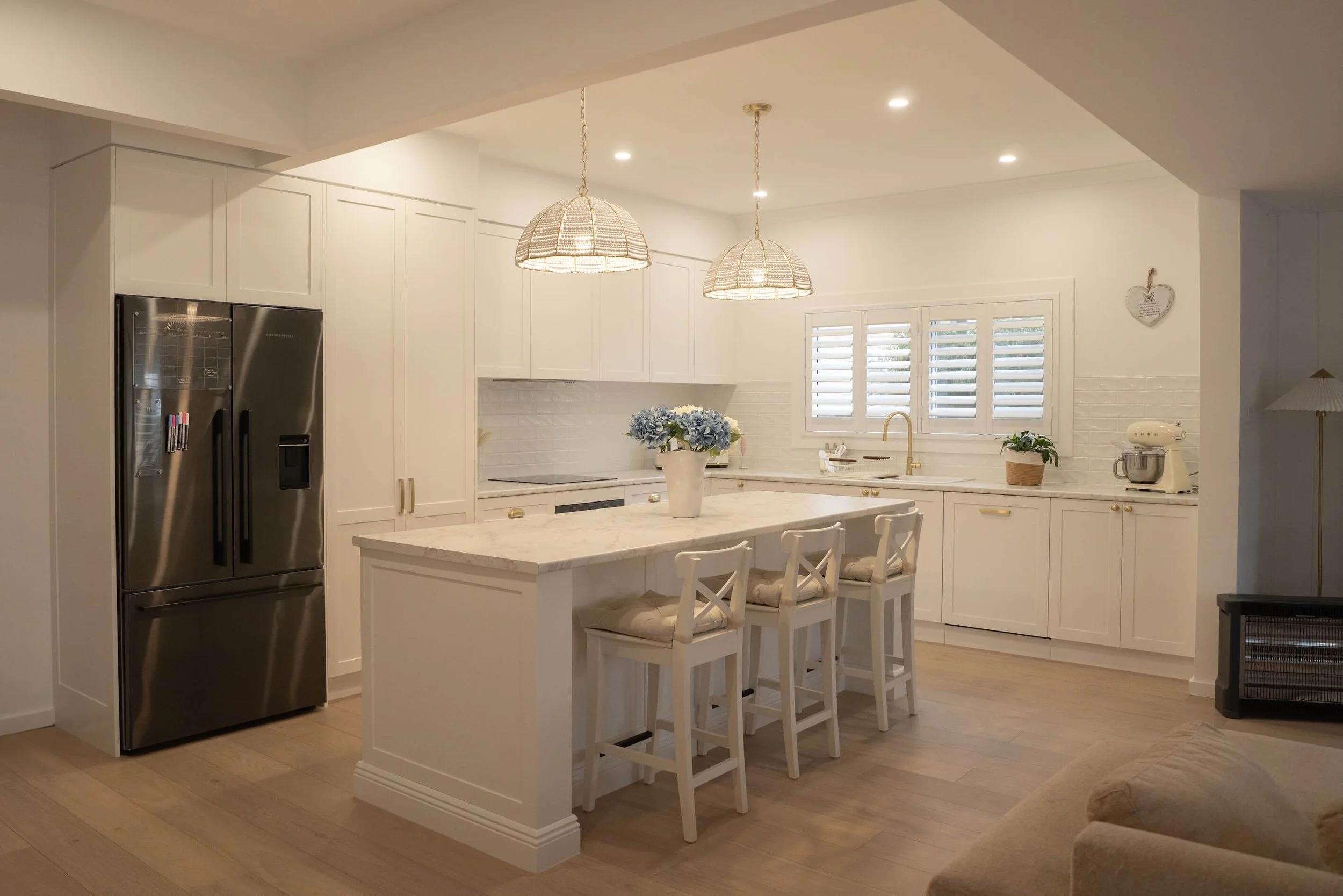 Bright, white kitchen with island, blue flowers, beige cushions on chairs, and window with shutters.
