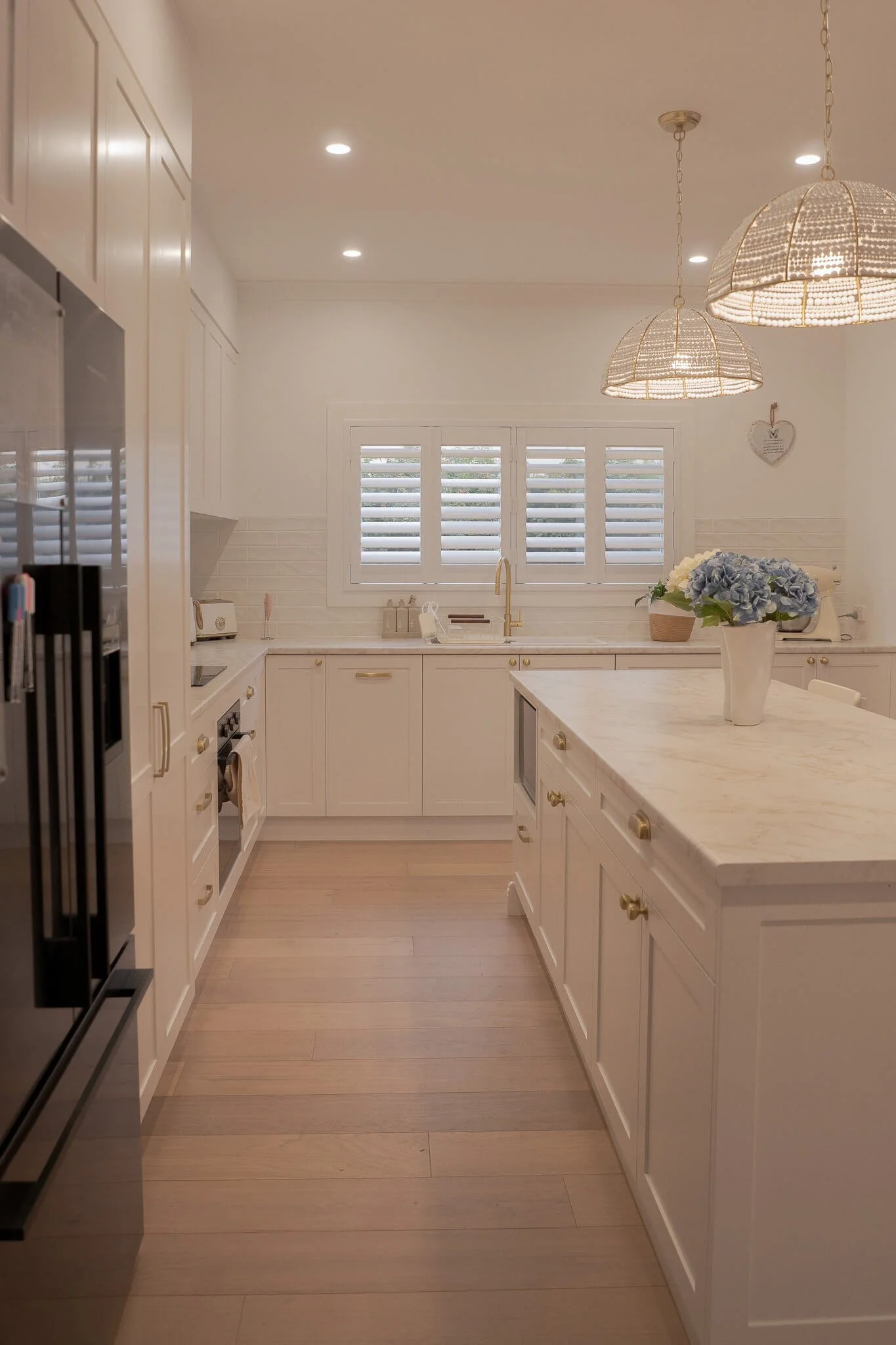 Bright white kitchen with two pendant lights, wooden floors, white cabinets, a window with shutters, and a counter with a vase of blue flowers.