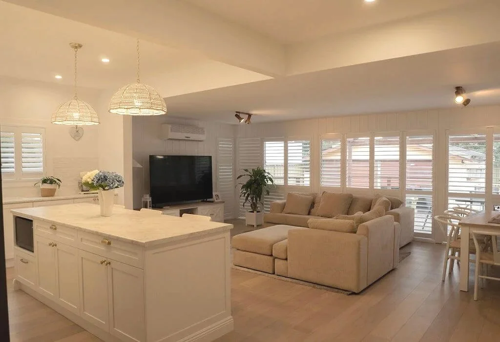 Bright, airy living room with a large L-shaped beige sofa, multiple windows with shutters, a flat-screen TV, a kitchen island with a flower vase, and hanging pendant lights.