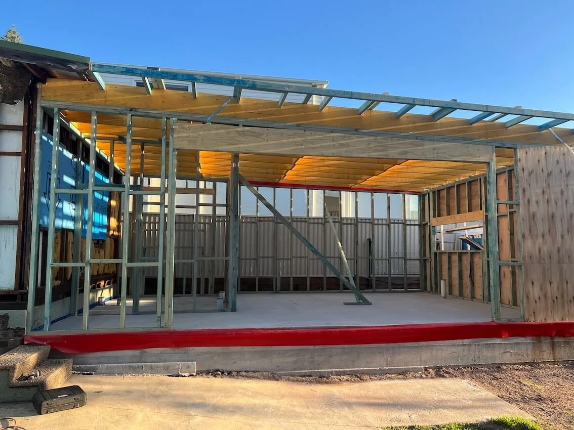 Construction of a building with exposed wooden and metal framing, concrete floor, and a partially finished roof under a clear blue sky.