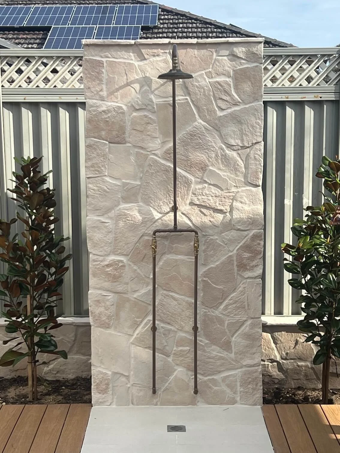Outdoor shower with black showerhead mounted on a beige stone wall, pipe extending downward, on a tiled patio with two small bushes on either side, metal fence and solar panels visible in the background.
