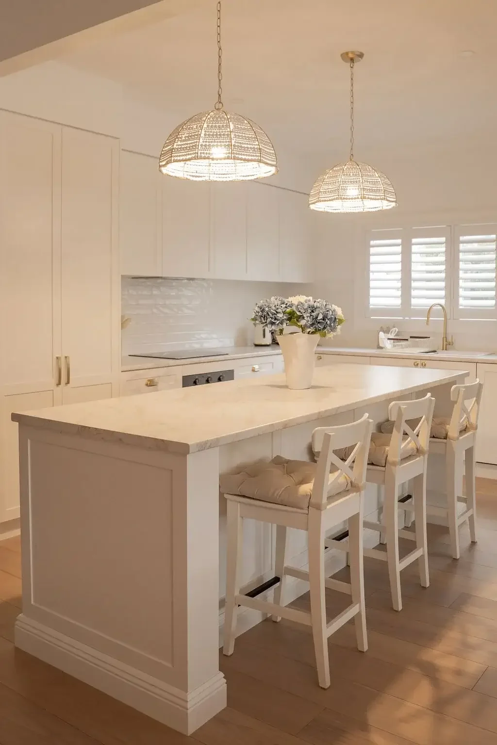 Bright, white kitchen with a large island, four white chairs with cushions, a white flower vase, and hanging pendant lights.