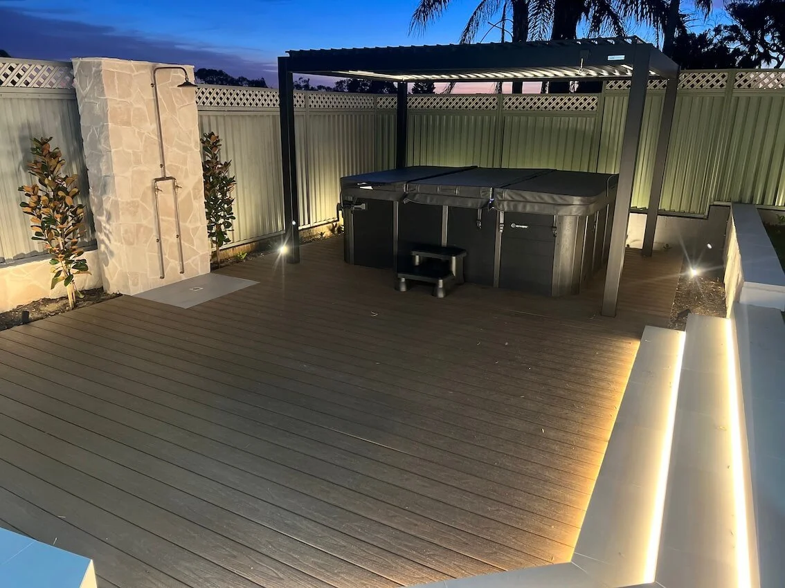 Outdoor deck area at dusk with a hot tub under a metal pergola, surrounded by a fence and small garden plants, illuminated by outdoor lighting.