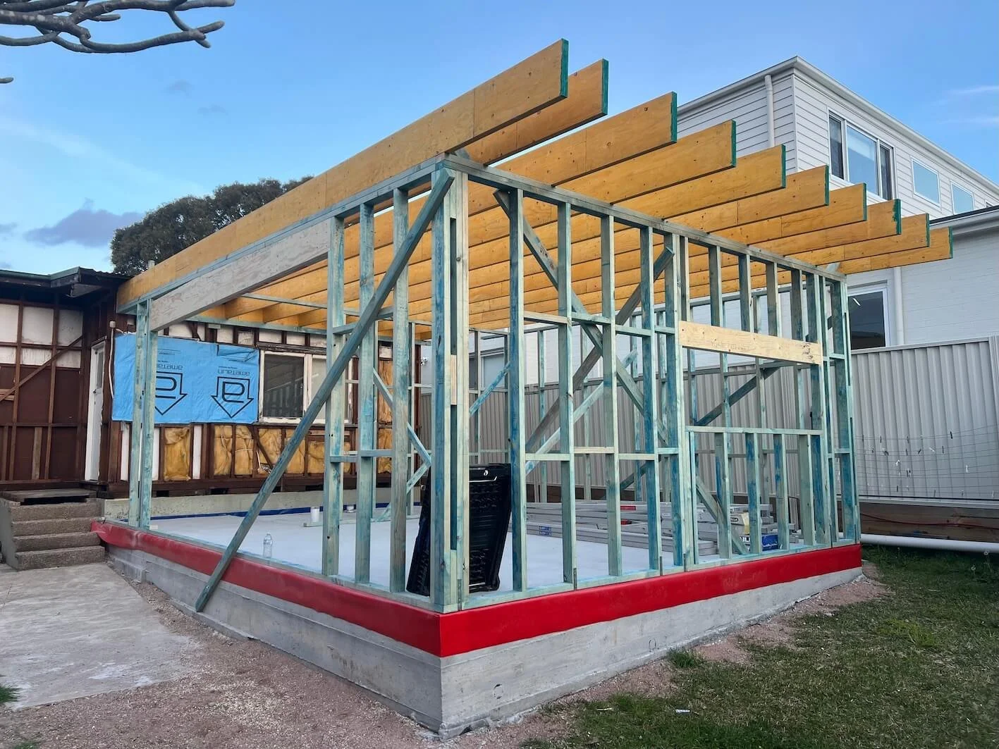 Construction site of a wooden deck and roof frame attached to a house, with framing in place and some roofing materials visible.