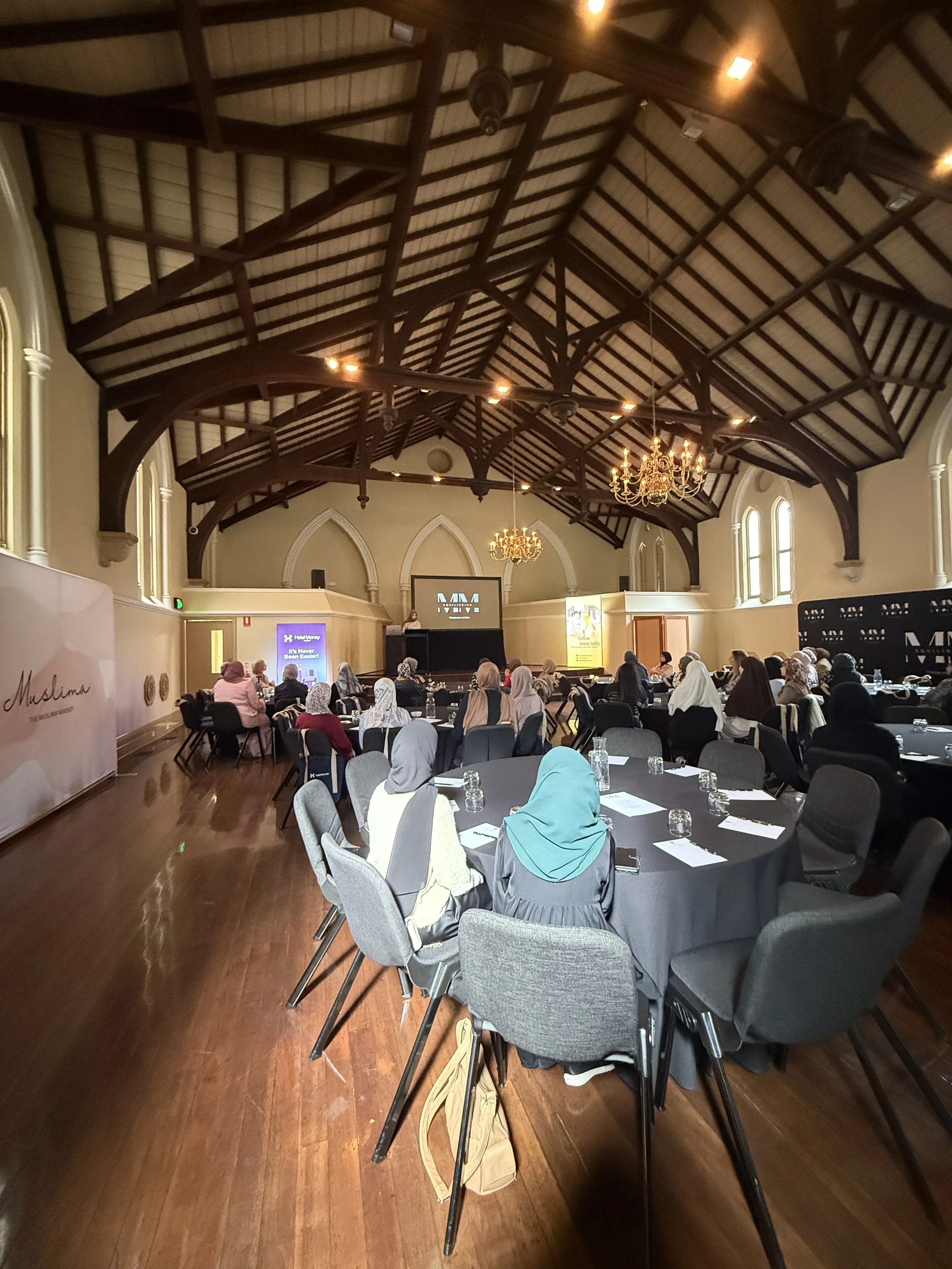 A conference room filled with women wearing headscarves, seated at round tables, facing a stage with a large screen and banners on the sides. The room has a high, wooden vaulted ceiling with chandeliers and is well-lit.