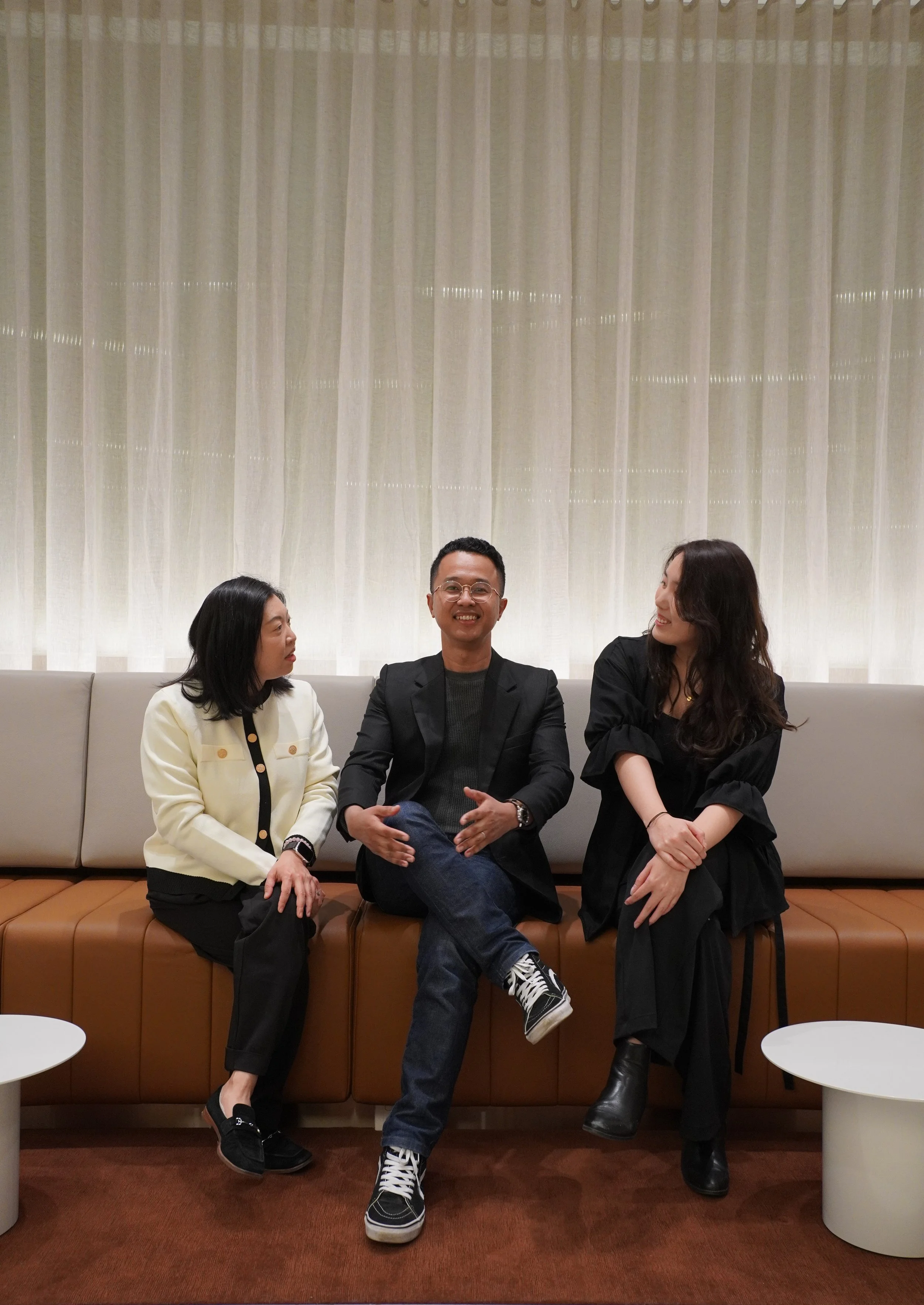 Three people sitting on a beige and brown sofa with a cream-colored curtain in the background. The man in the middle is smiling, wearing glasses, a black blazer, and jeans. The woman on the left is wearing a cream-colored jacket and black pants, and the woman on the right is dressed in black, with long wavy hair and black boots.