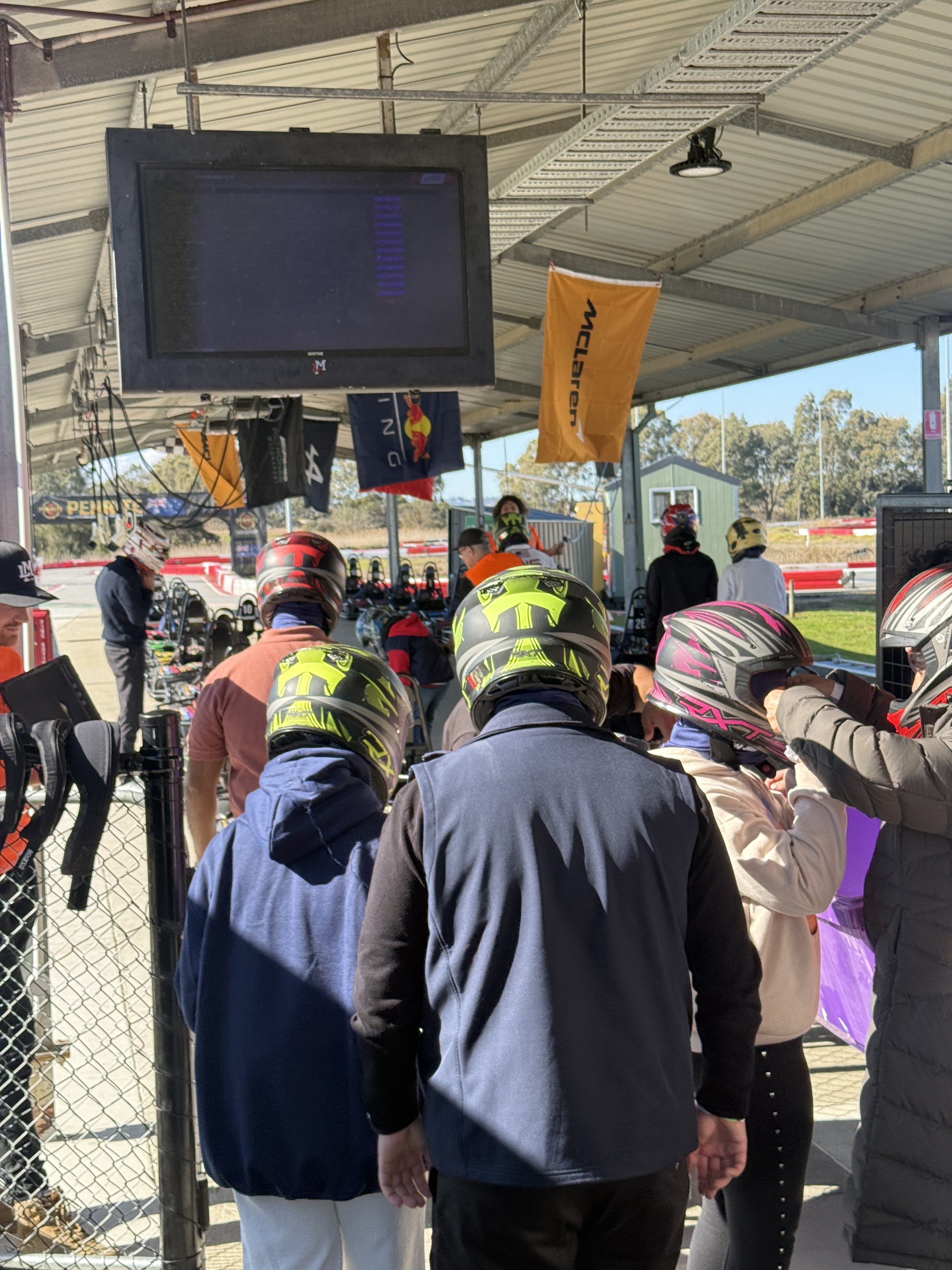 People wearing motorcycle helmets and jackets gathered at a race track clubhouse, with flags and a large screen above.