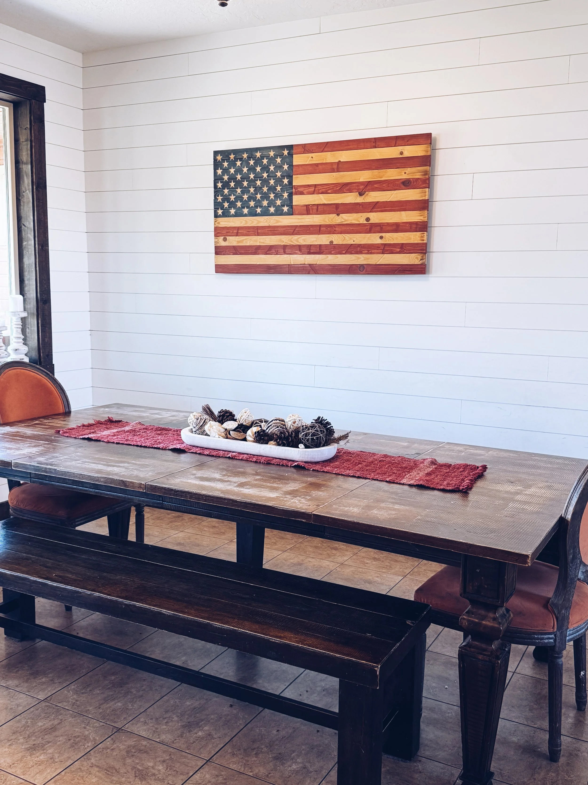 A dining room with a wooden table, a red runner, and a centerpiece of pinecones and decorative balls. On the wall is a wooden American flag, and there are chairs and a bench around the table.