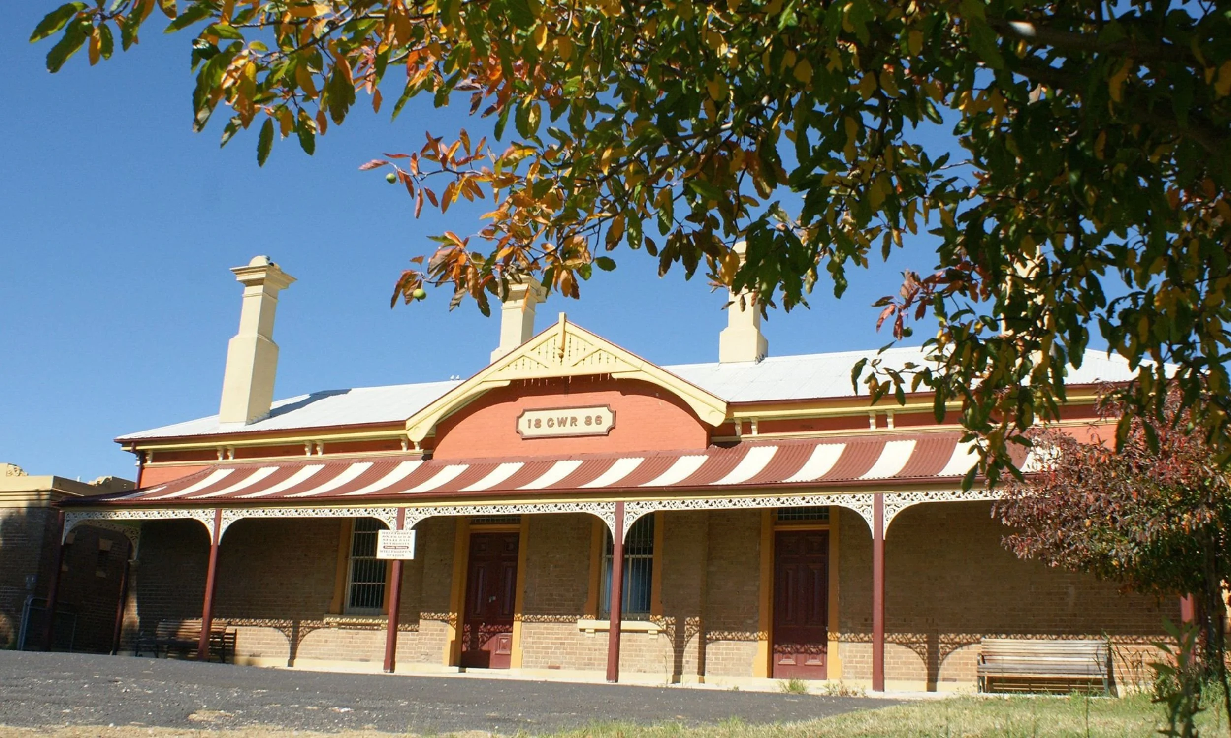 Historical brick building with a decorative metal awning, a sign displaying '18 GWR 86,' three chimneys on the roof, and trees with autumn leaves partly framing the image under a clear blue sky.