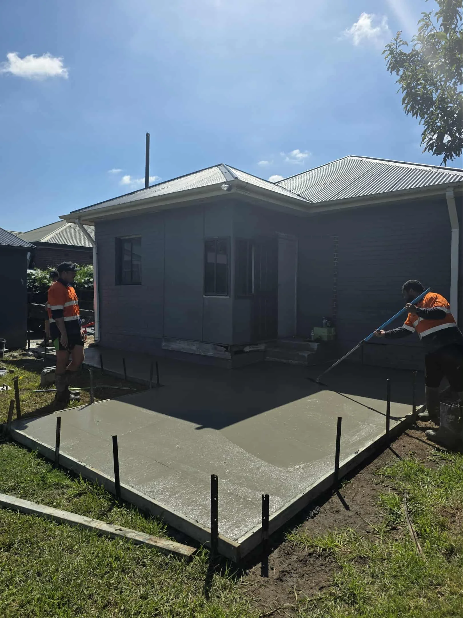 Two construction workers smooth wet concrete in a small backyard patio area in front of a house.