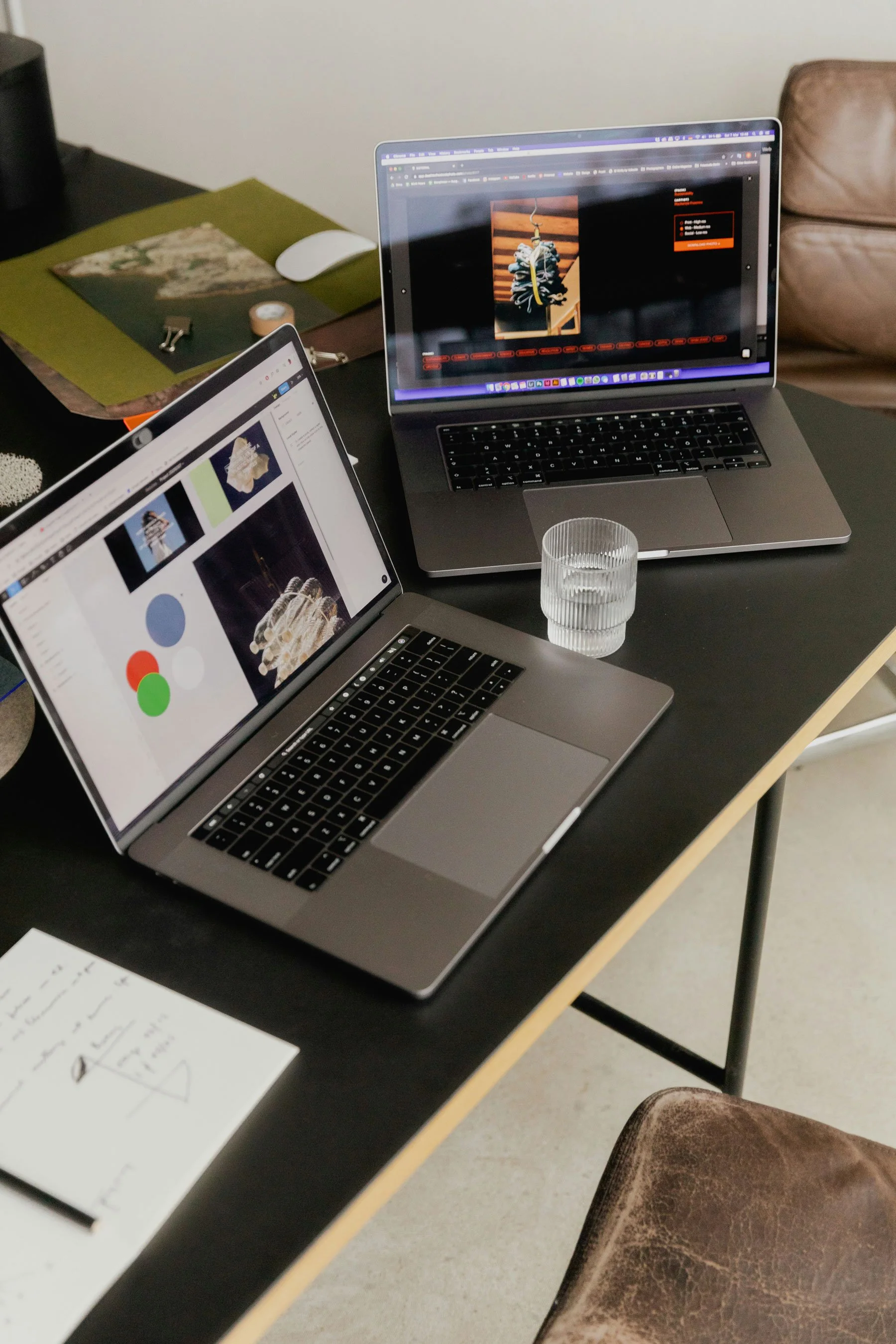 Two open laptops on a black table, one displaying a digital art project and the other a webpage, with a glass of water, a notepad, and office supplies nearby.