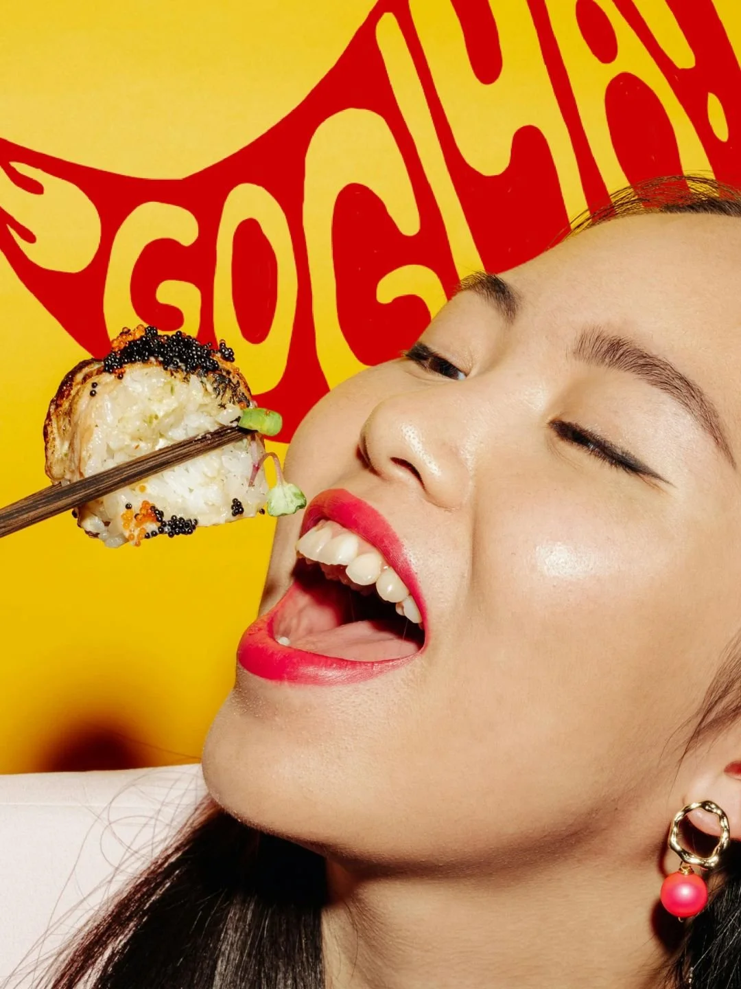A woman with red lipstick and earrings opening her mouth while a piece of sushi is about to be placed inside with chopsticks, standing in front of a yellow background with red writing.