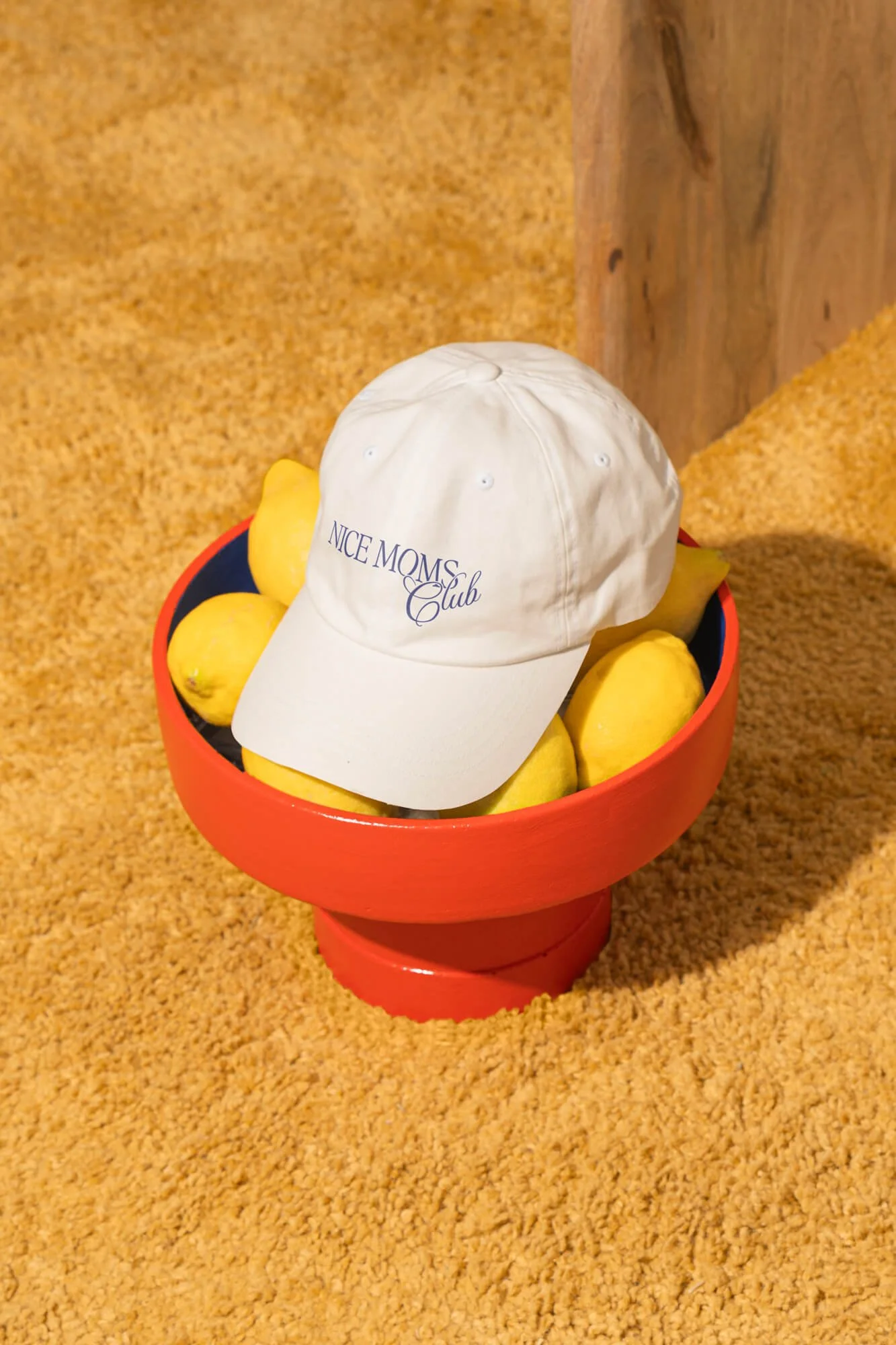 A red bowl filled with lemons and a white baseball cap with the words 'Nice Moms Club' embroidered on it, placed on a yellow carpeted floor.