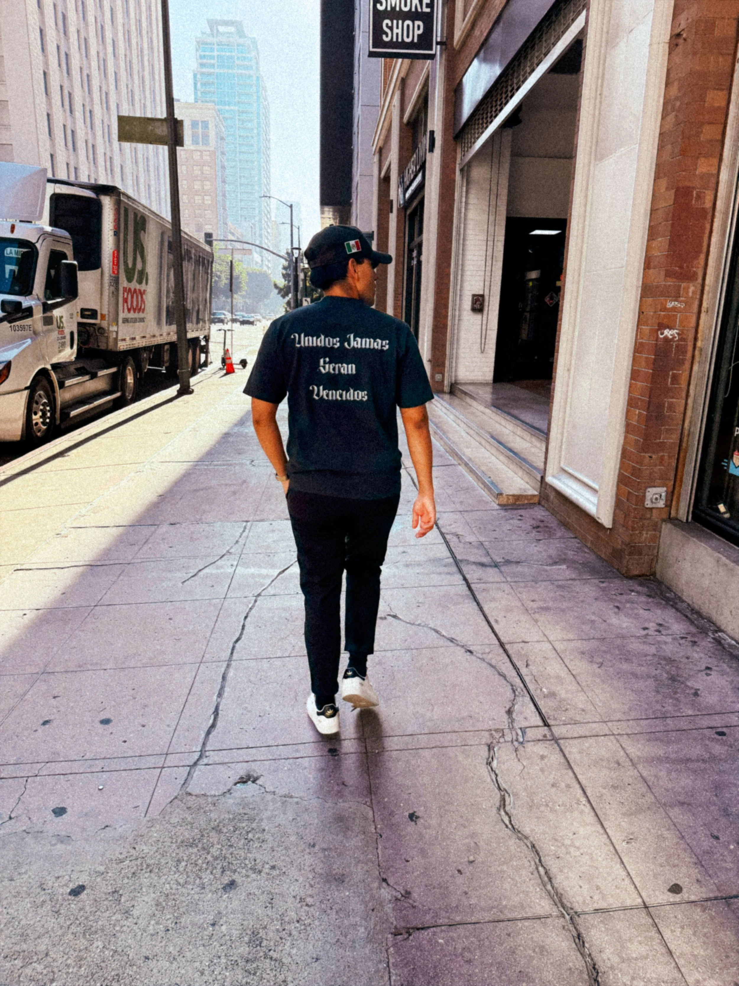 Person walking on city sidewalk, wearing black cap and shirt with Spanish text, near storefronts and parked trucks.