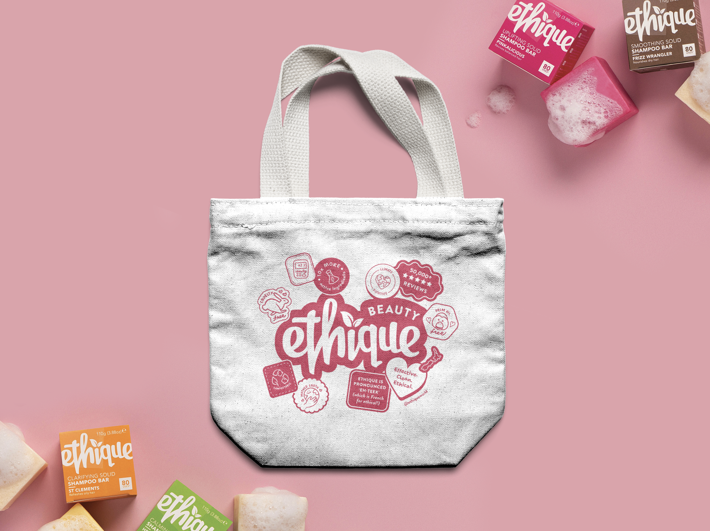 Gray tote bag with pink branding and text, surrounded by soap bars and shampoo boxes on a pink background.
