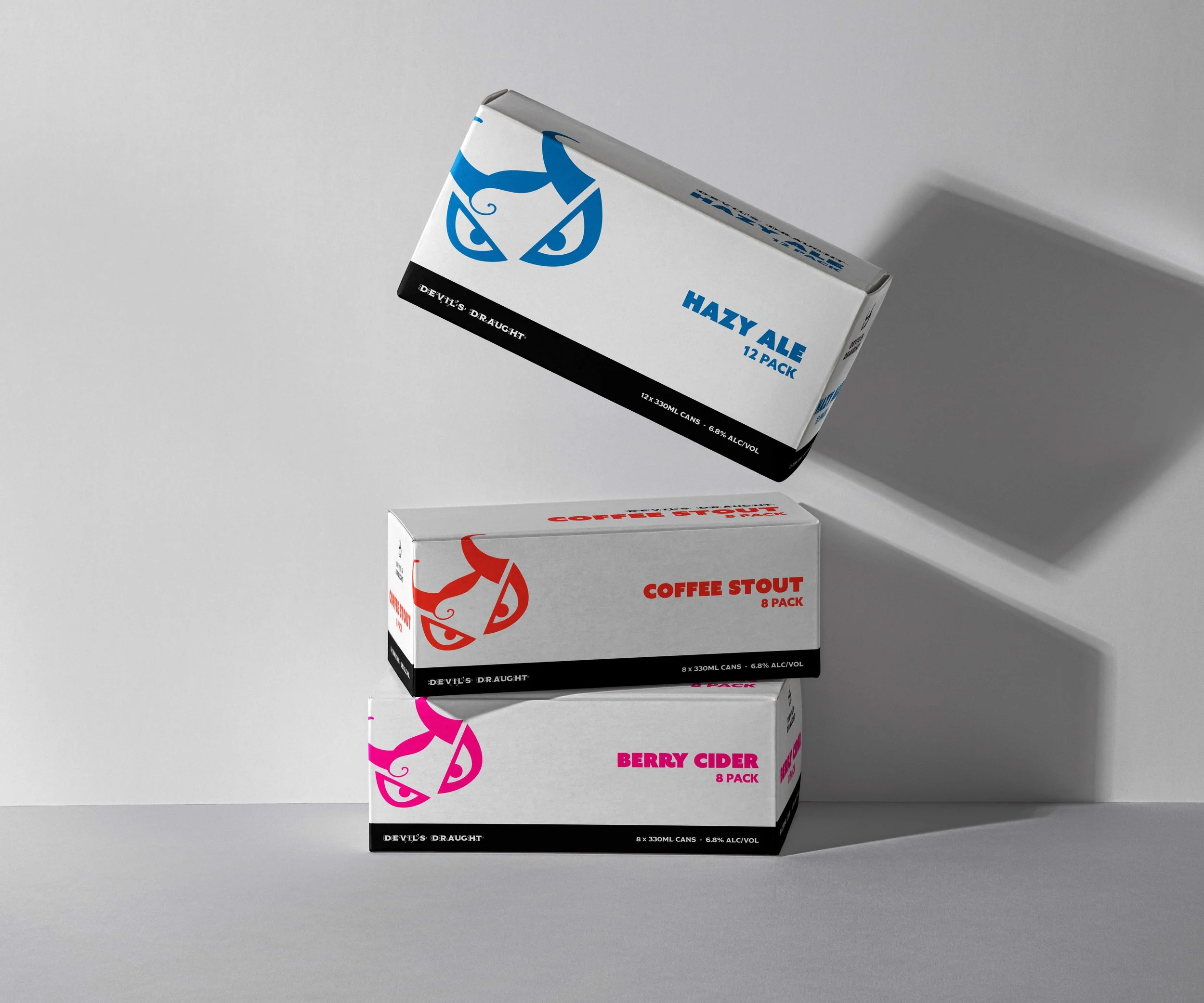Three stacked beverage boxes labeled Hazy Ale, Coffee Stout, and Berry Cider from Devil's Draught company, with each logo featuring a stylized devil's face in blue, red, and pink respectively, casting shadows on a plain background.