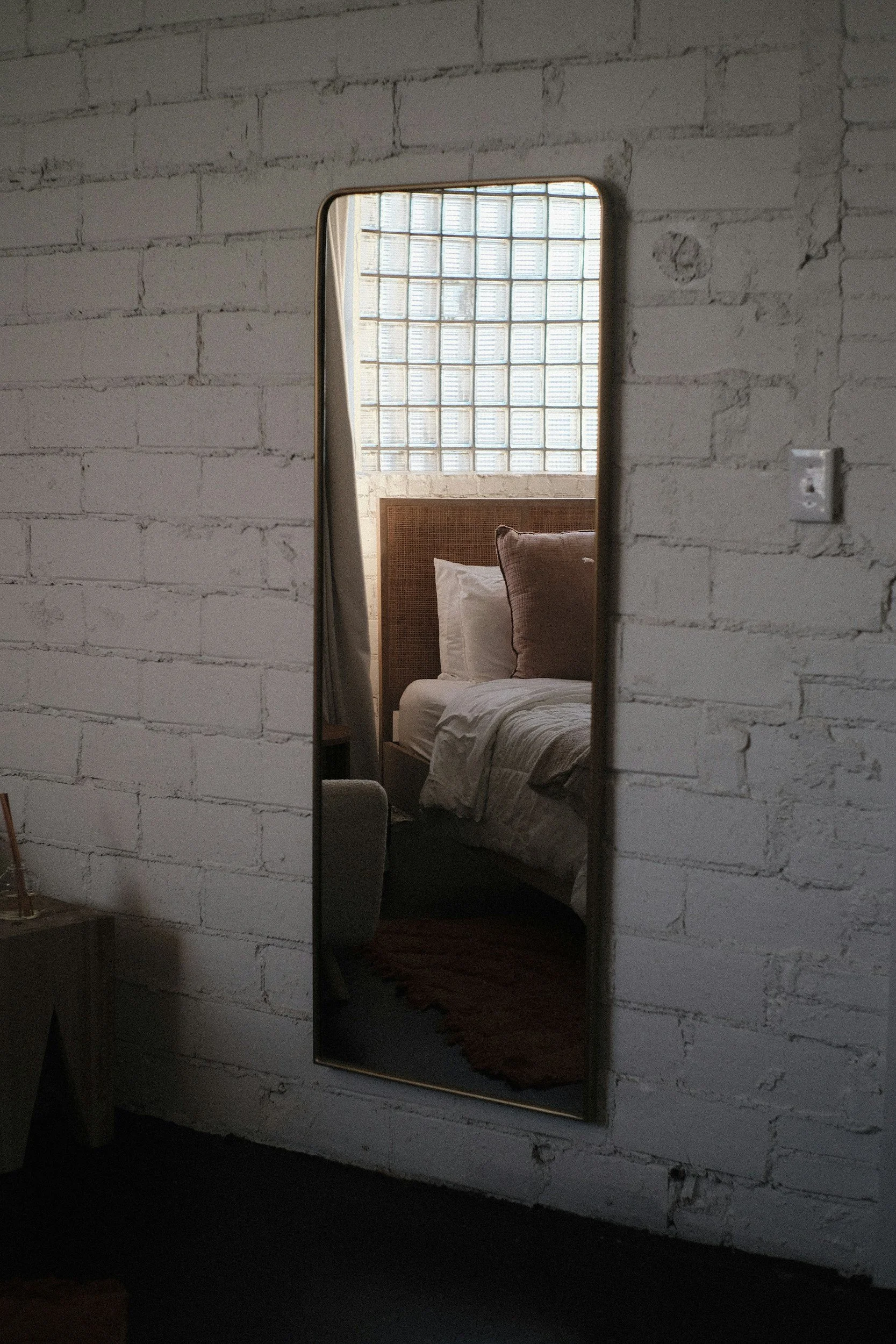 Reflection of a bed with pillows and a headboard, seen in a tall mirror mounted on a white brick wall in a room with a glass block window.