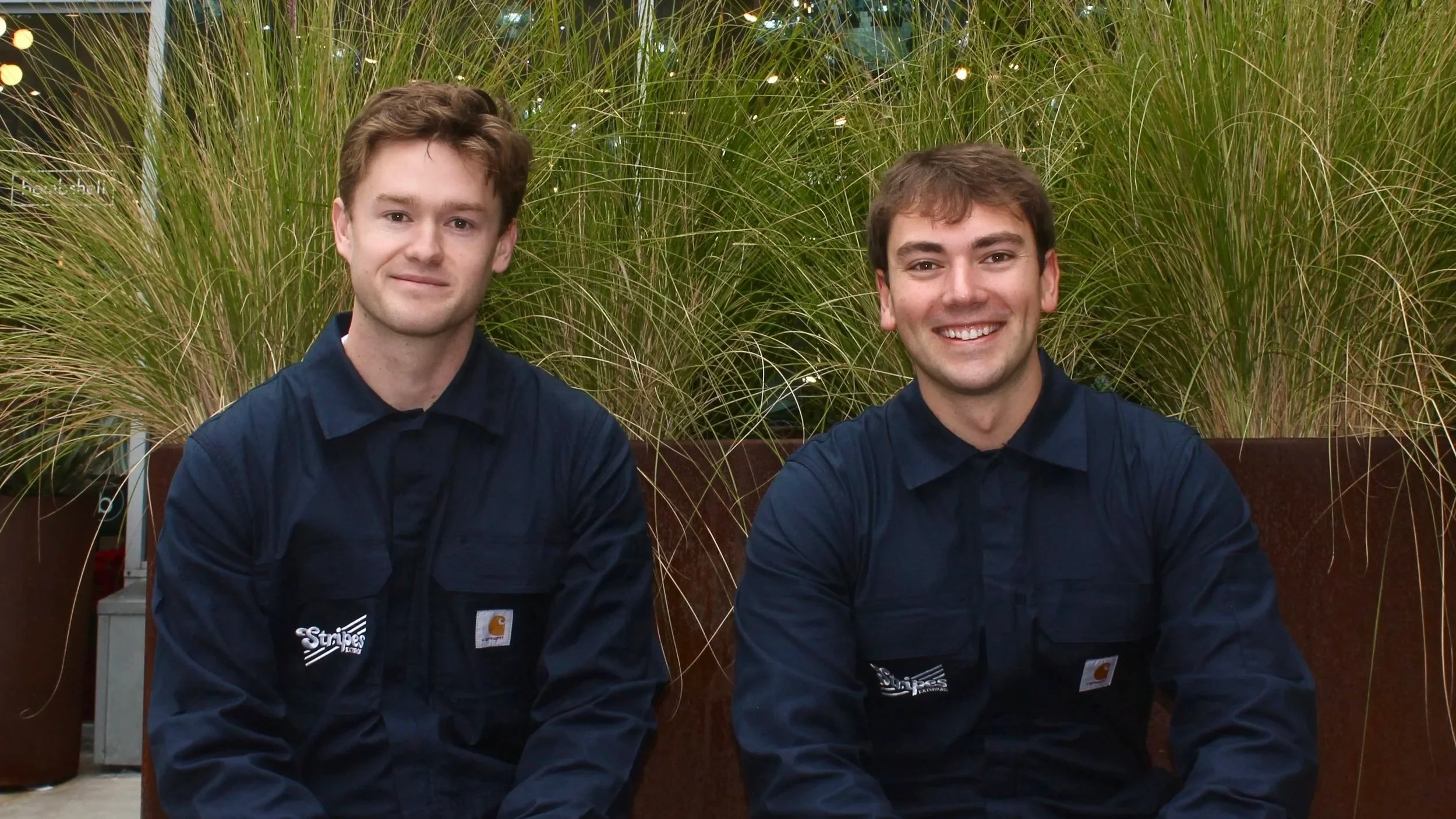 Two young men wearing dark blue work shirts with logos, sitting in front of tall green ornamental grass plants inside a building.