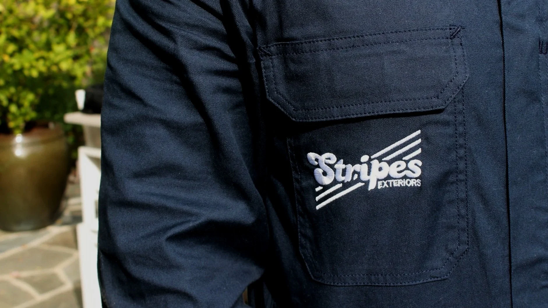 Close-up of a dark navy work jacket with a pocket that has white embroidered text reading 'Stripes Exteriors' and a striped logo, outdoors with a potted plant and stone pavement in the background.