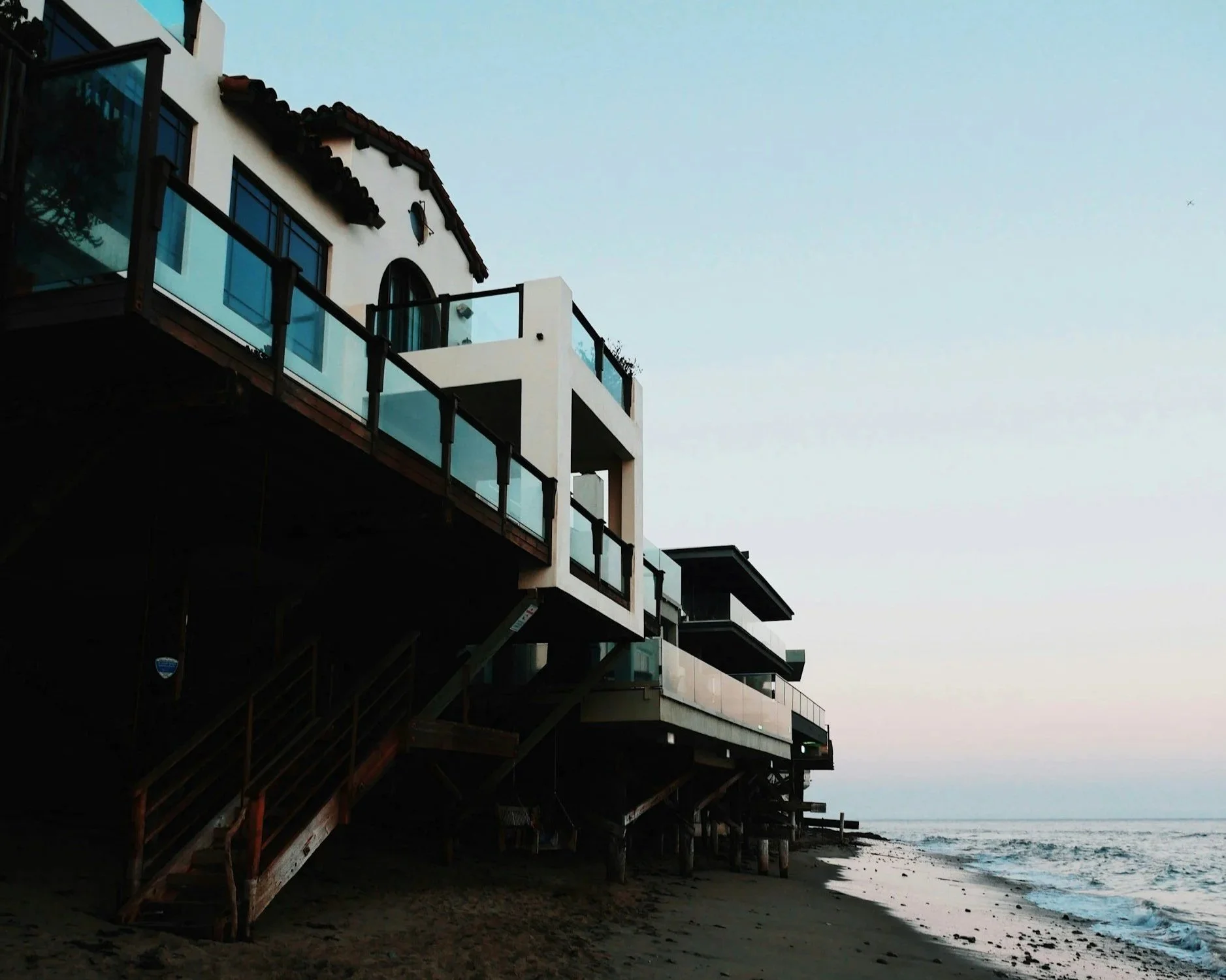 Beachfront houses on stilts with ocean waves, clear sky, and a sandy shore, during sunset or sunrise.