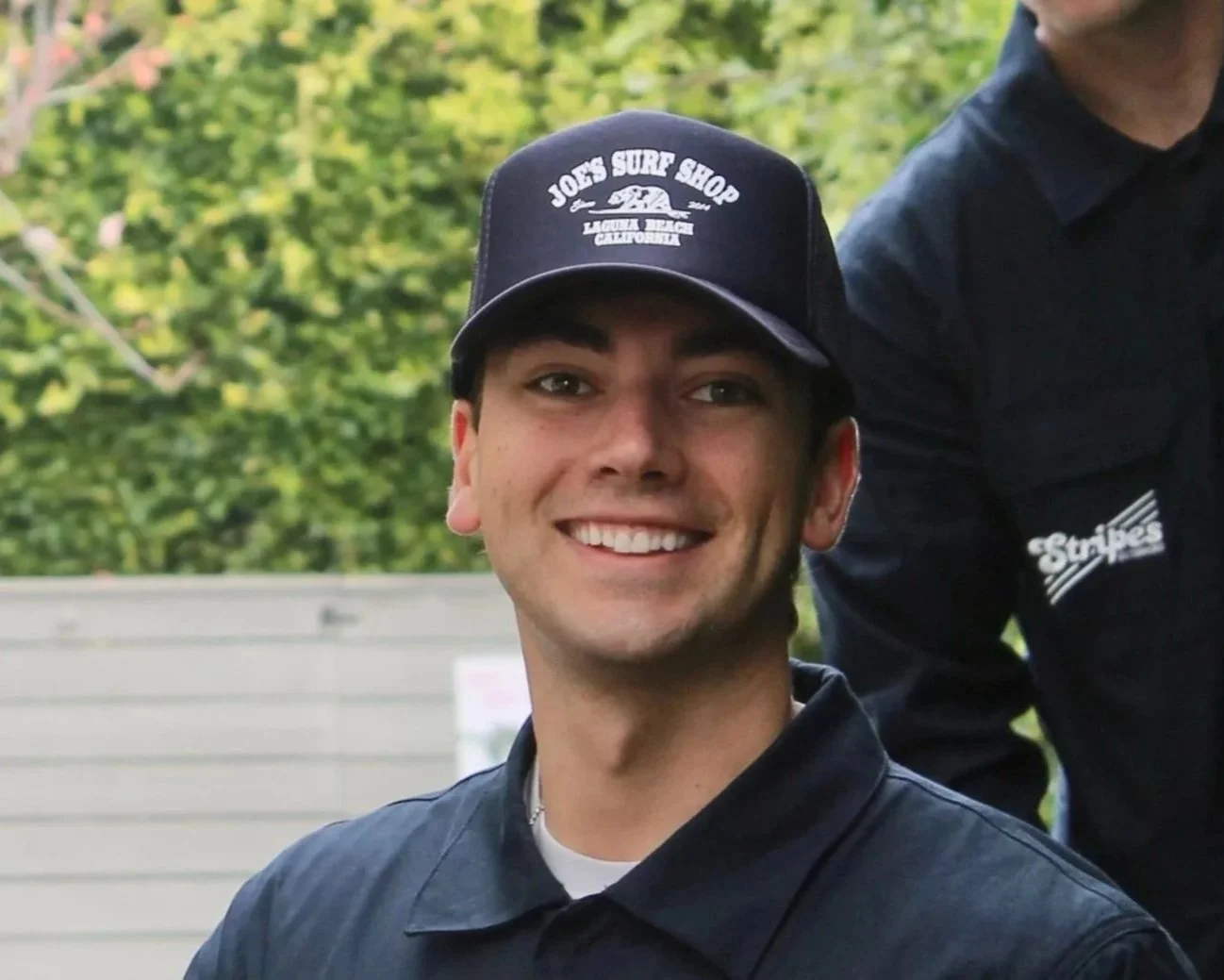 A smiling young man wearing a black cap with a logo and text, and a black polo shirt, outdoors with greenery in the background.