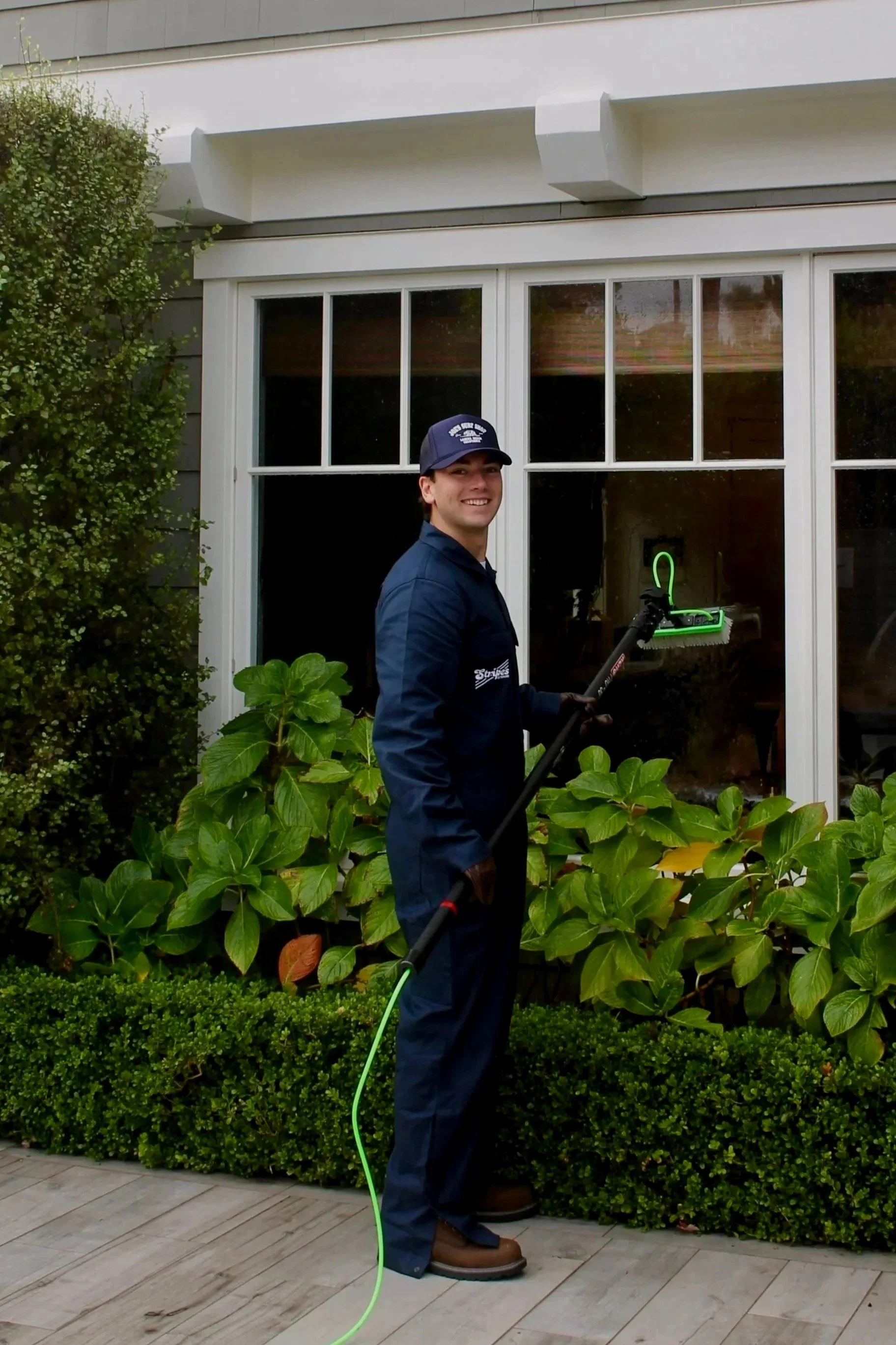 A man in navy work overalls and a cap is using a window cleaning tool to wash exterior windows of a house. The house has white-framed windows, gray siding, and there are green bushes and plants below the windows.