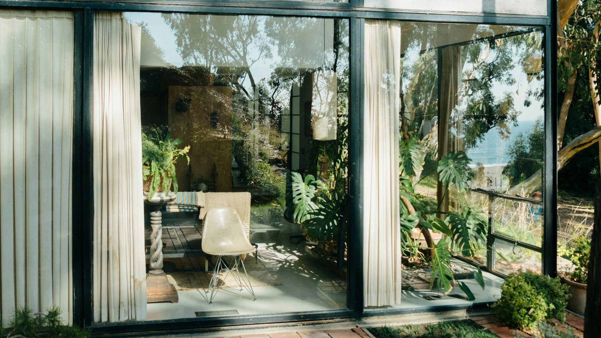 View through a large glass window of an indoor space with white curtains, a beige chair, a small table with a plant, and lush green plants and trees outside.