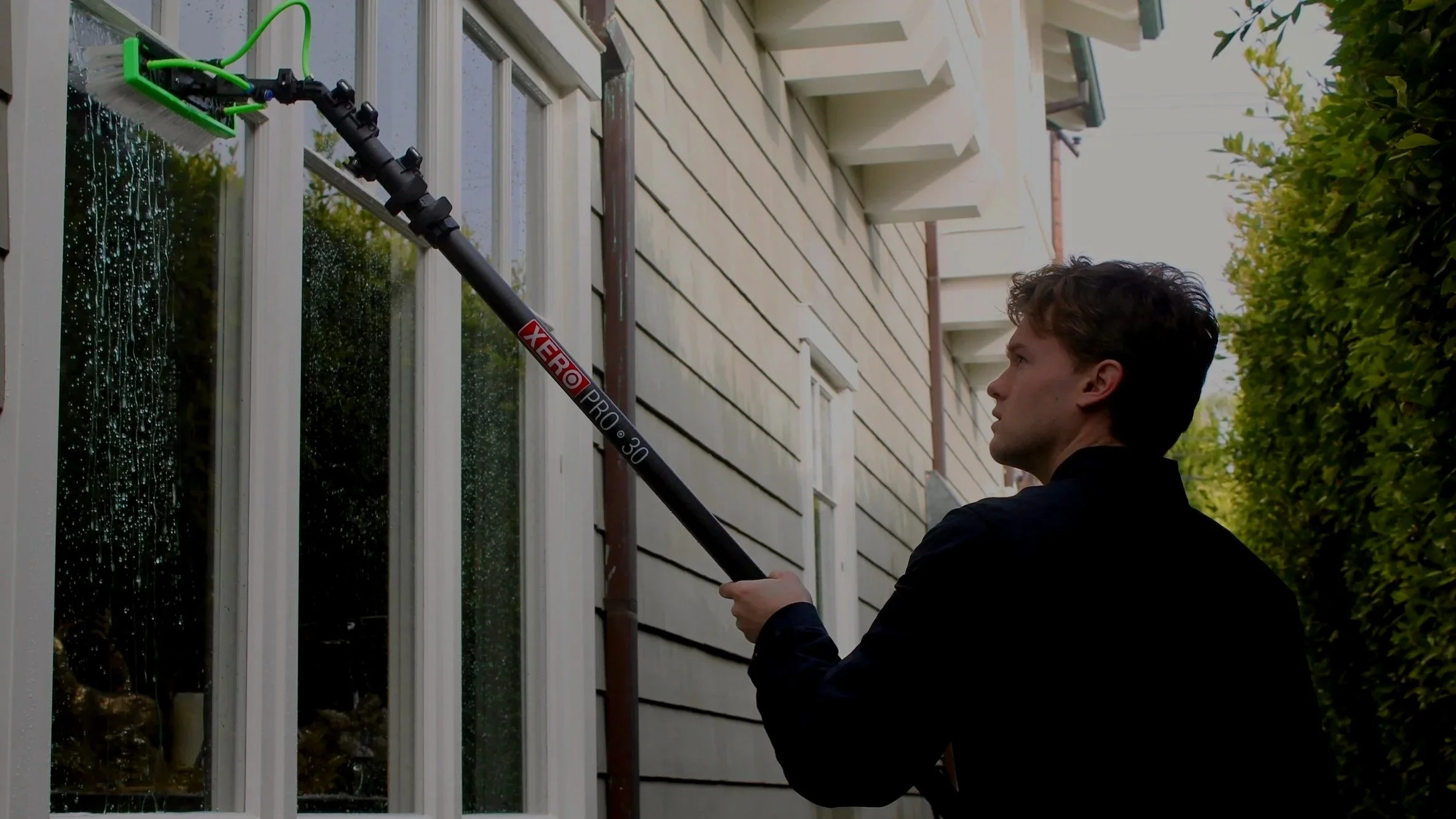 A man using a long extension pole with a brush attachment to clean or wash a window on the exterior of a house on a sunny day.