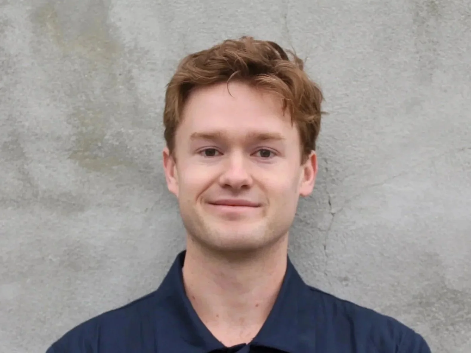 A young man with red hair and fair skin smiling in front of a plain, gray concrete wall.