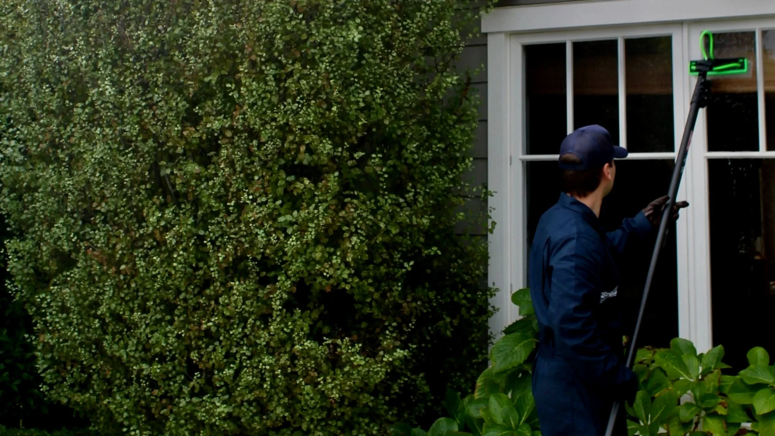 A person wearing a navy uniform and cap using a long-handled cleaning tool to wash a window in a house with a gray siding exterior. There are green bushes and plants nearby.