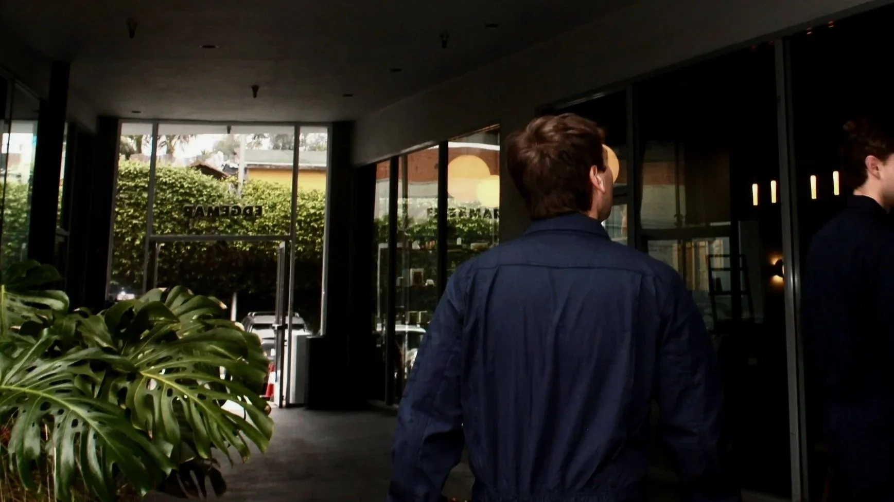 A person wearing a dark blue shirt walking into a building with glass doors, with large green leafy plants on the left side and outside through the glass, trees and vehicles are visible.