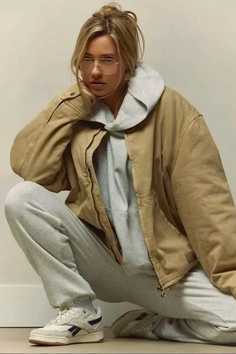 Person squatting down, wearing glasses, a tan jacket over a gray hoodie, gray sweatpants, and white sneakers, with a contemplative expression.