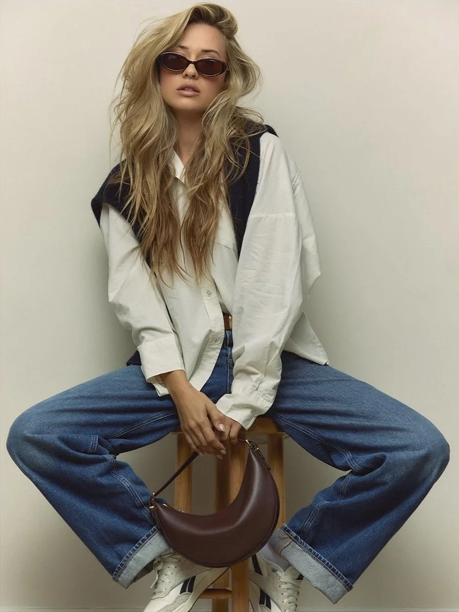 A woman with long, wavy blonde hair wearing sunglasses, a white oversized shirt, blue jeans, and white sneakers, sitting on a wooden stool against a plain, light-colored wall, holding a brown crescent-shaped handbag.