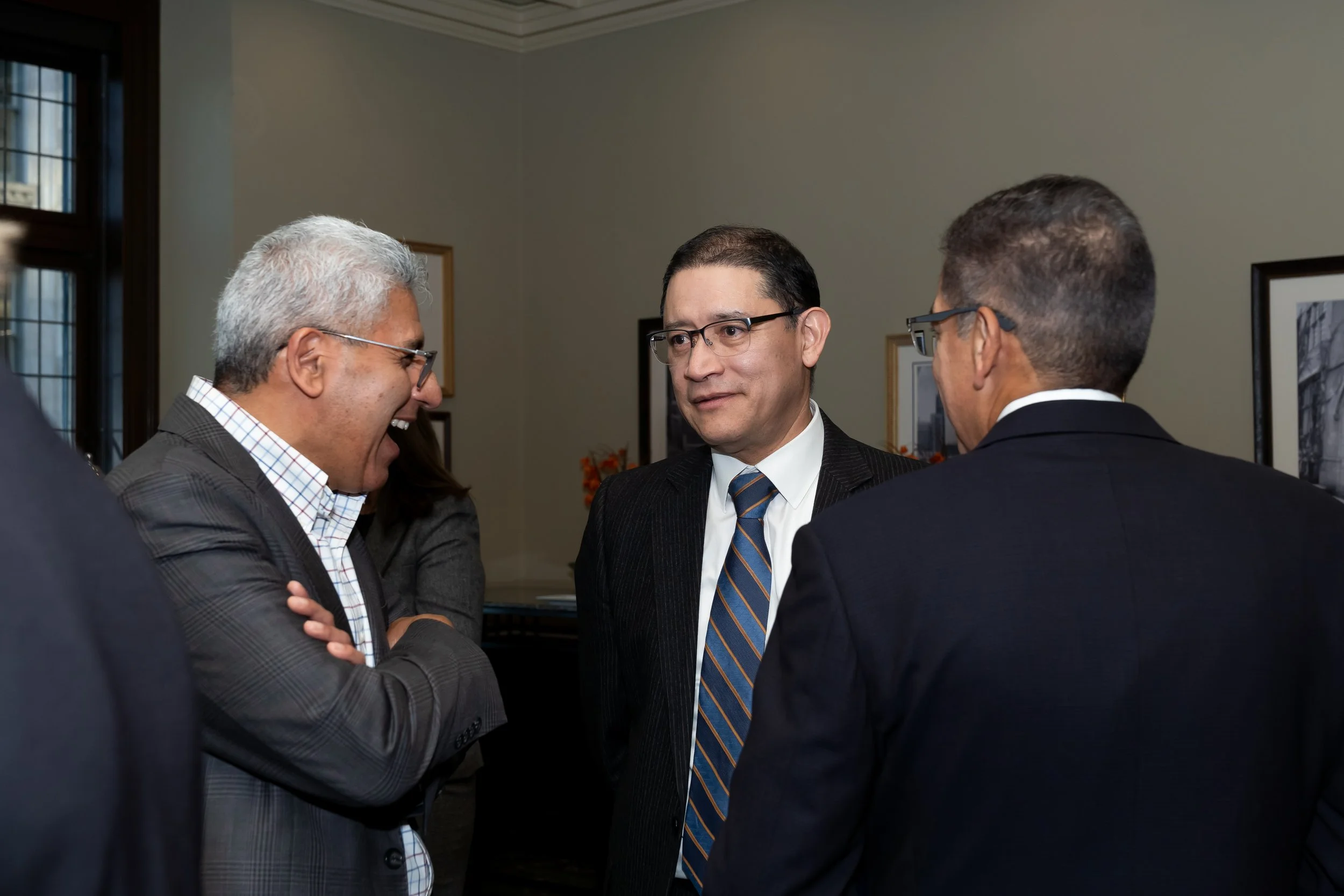 Three men in business suits having a conversation in a room with framed pictures on the wall.