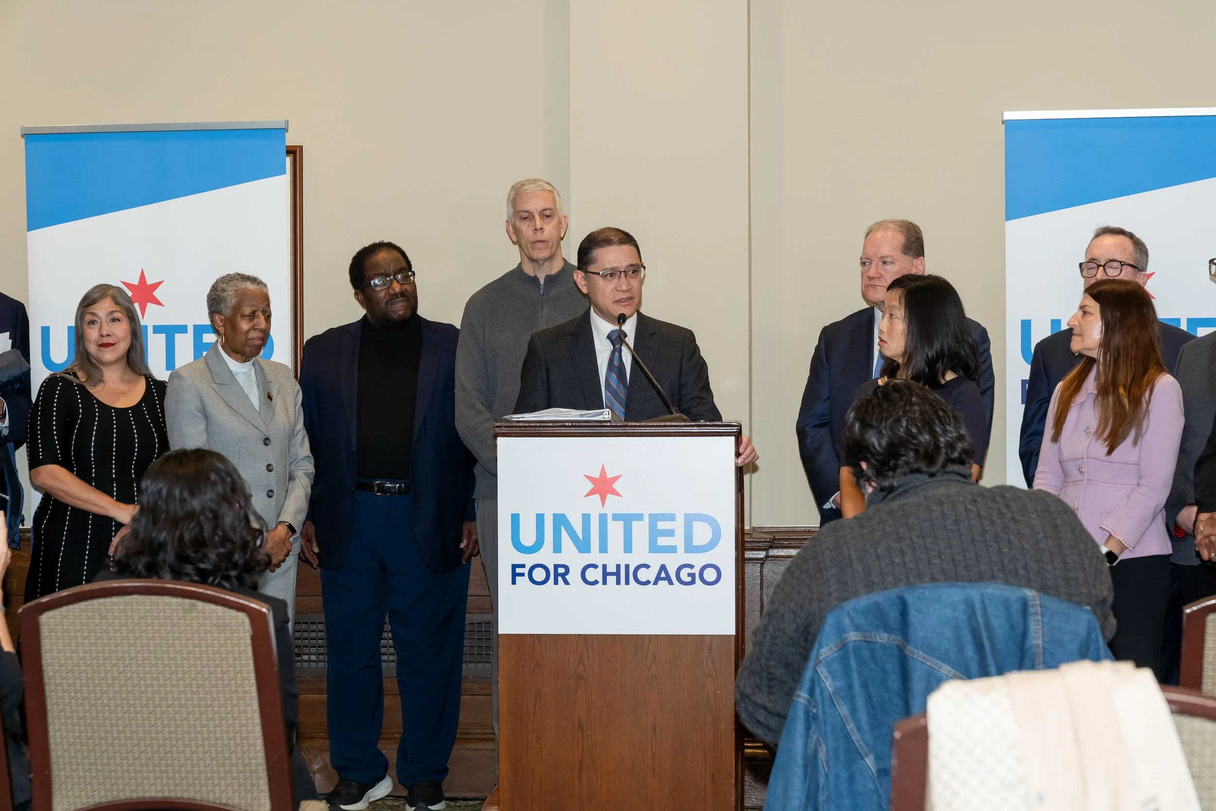 A group of diverse people standing behind a podium with a sign that reads 'United for Chicago' during a press conference or political event.