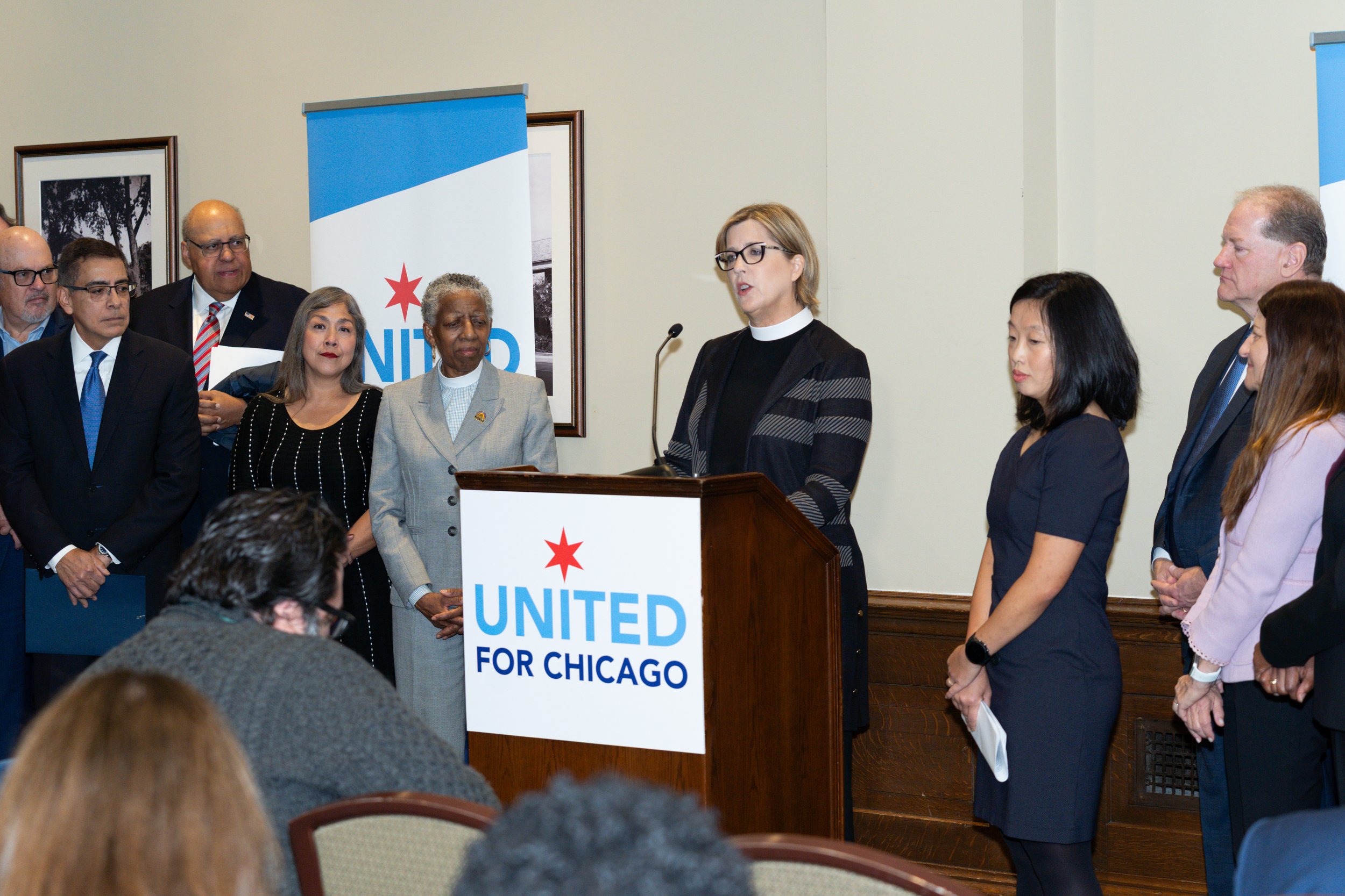 A woman speaking at a podium at a political event with a sign that reads 'United for Chicago.' Several people are standing behind her, listening attentively, in a room with framed pictures on the wall.