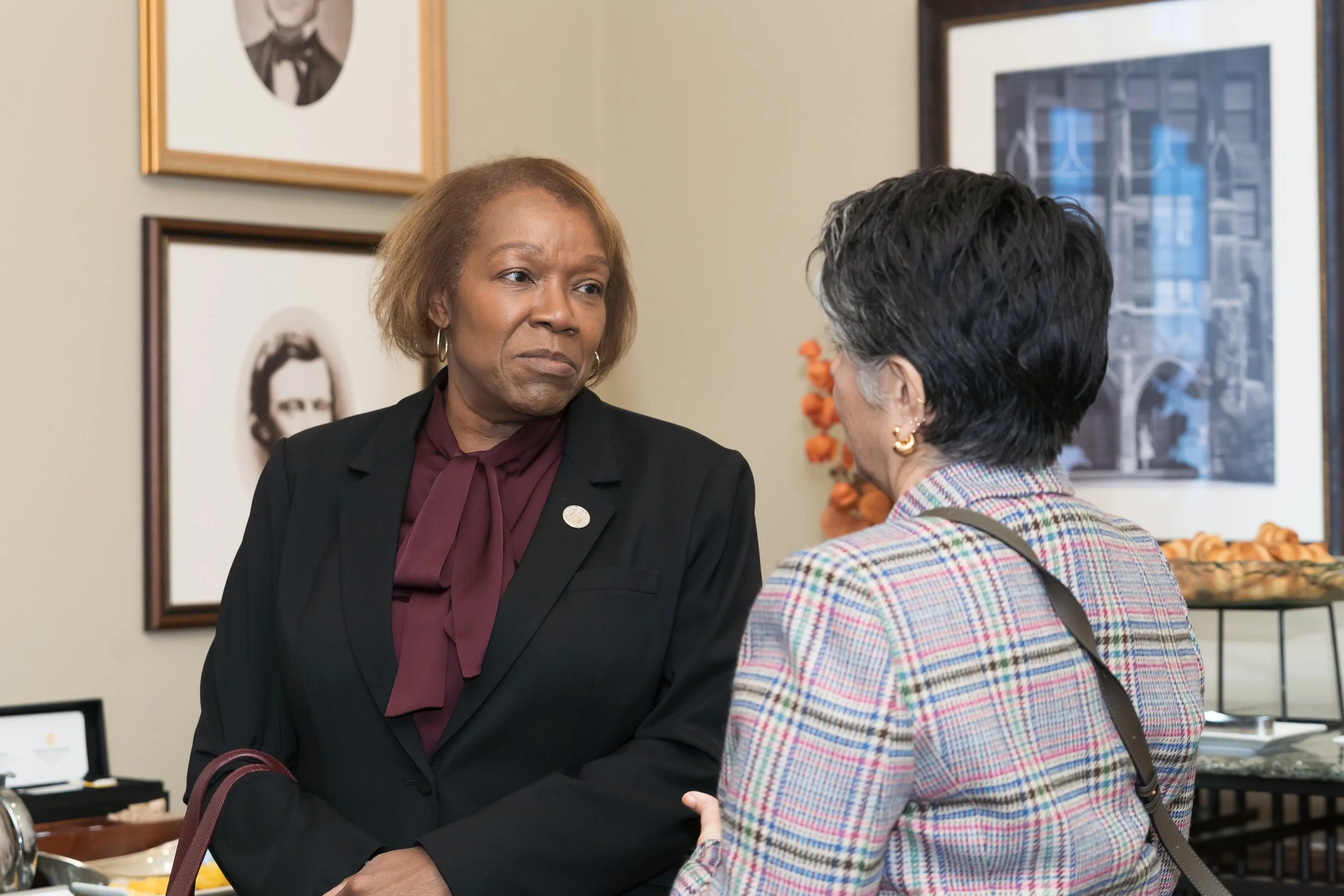 Two women engaged in a serious conversation indoors, with framed photographs and artwork on the wall behind them.
