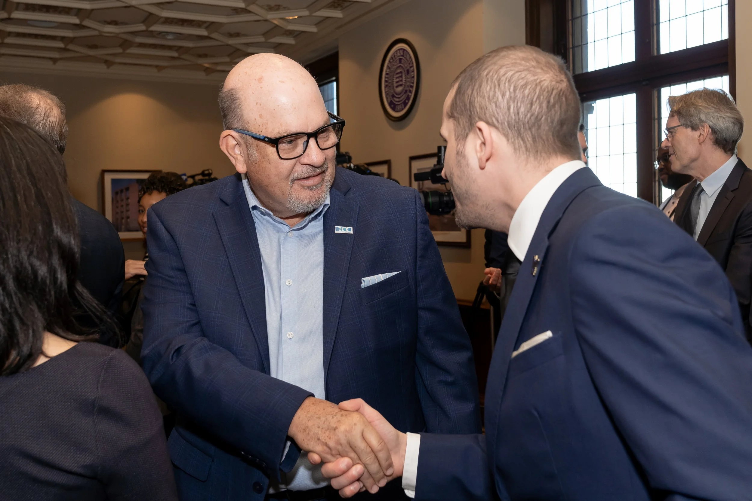 Two men in suits shaking hands in a professional setting, with people and cameras in the background.